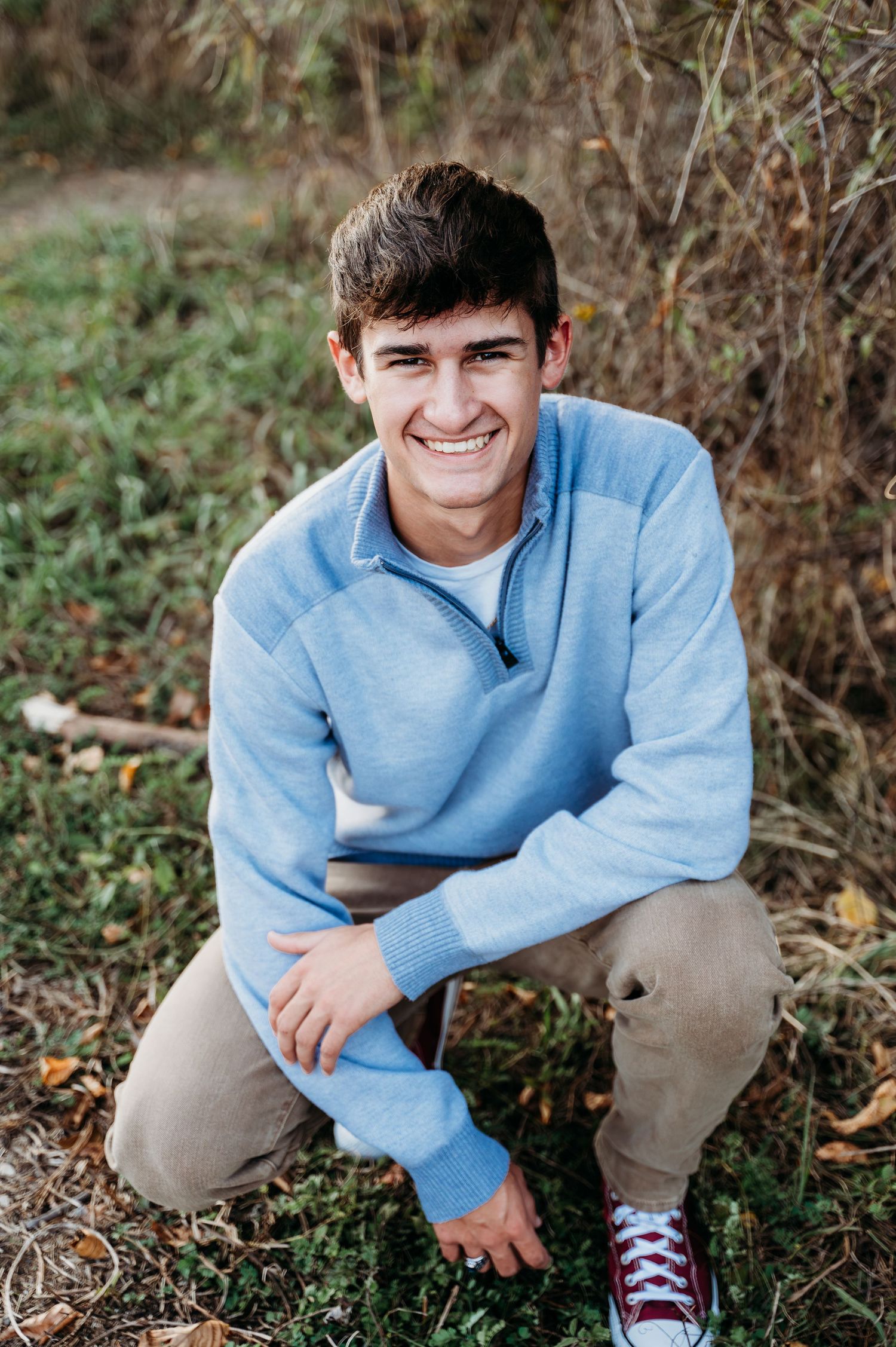 Person in light blue sweater and khaki pants crouches outdoors in casual autumn setting.