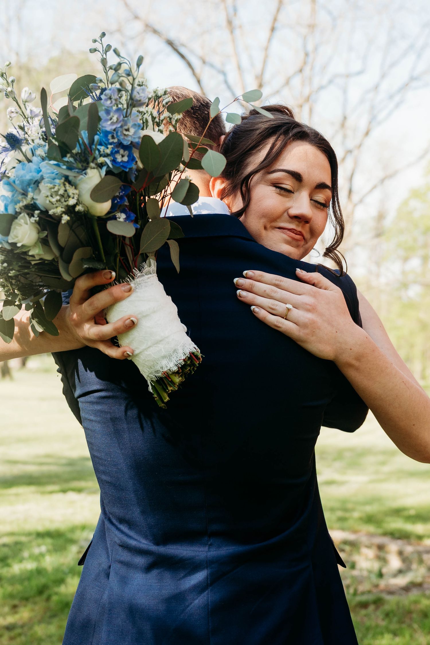 Bride smiling with eyes closed, hugging groom outdoors, holding blue and white wedding bouquet, wearing engagement ring.
