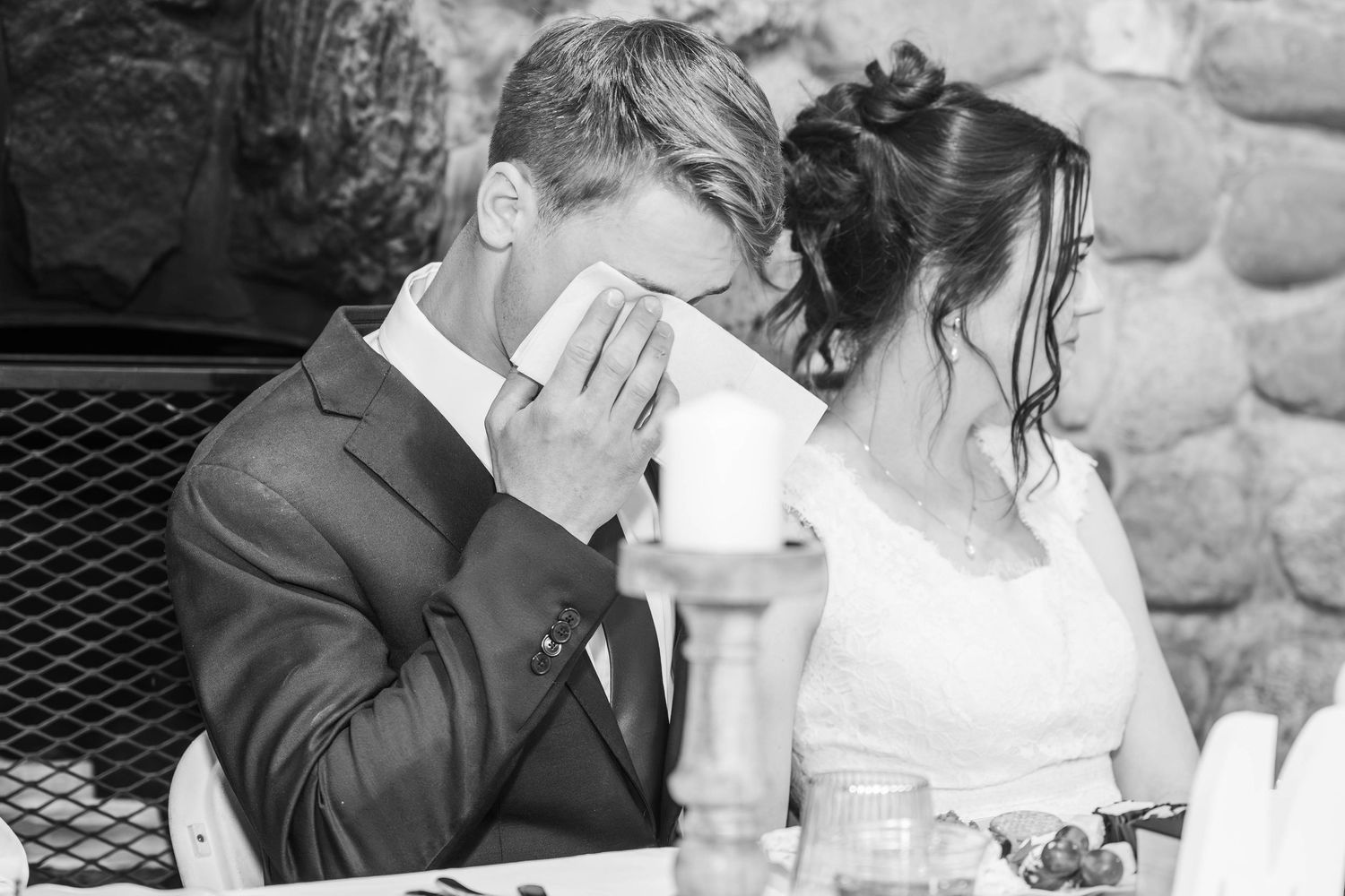 Black and white wedding photo of groom wiping tears beside bride at reception table.