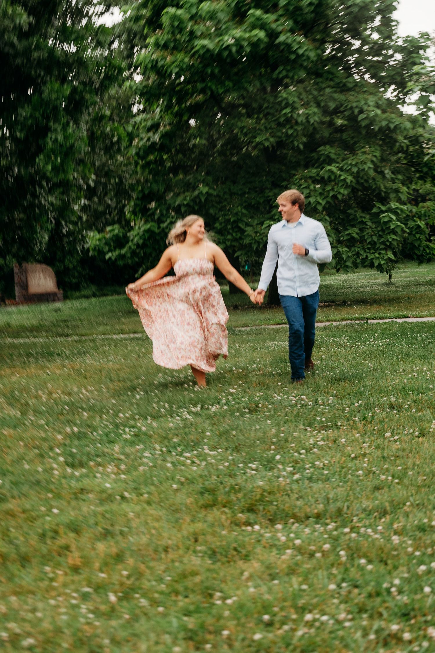 A couple holding hands and running joyfully through a lush green park, woman in a flowing pink floral dress.
