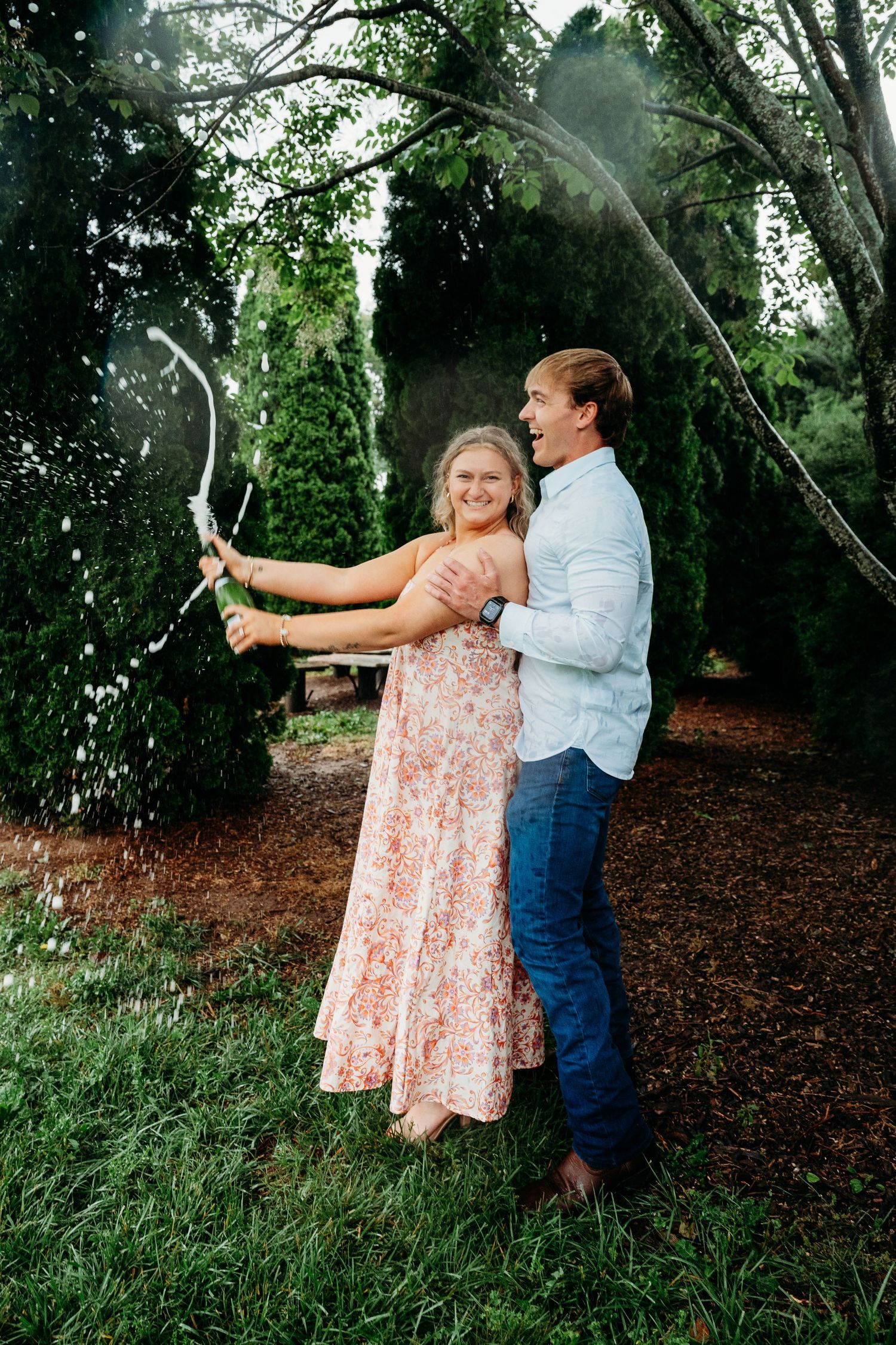 Joyful couple popping champagne bottle outdoors, woman in floral pink dress, man in light blue shirt and jeans.