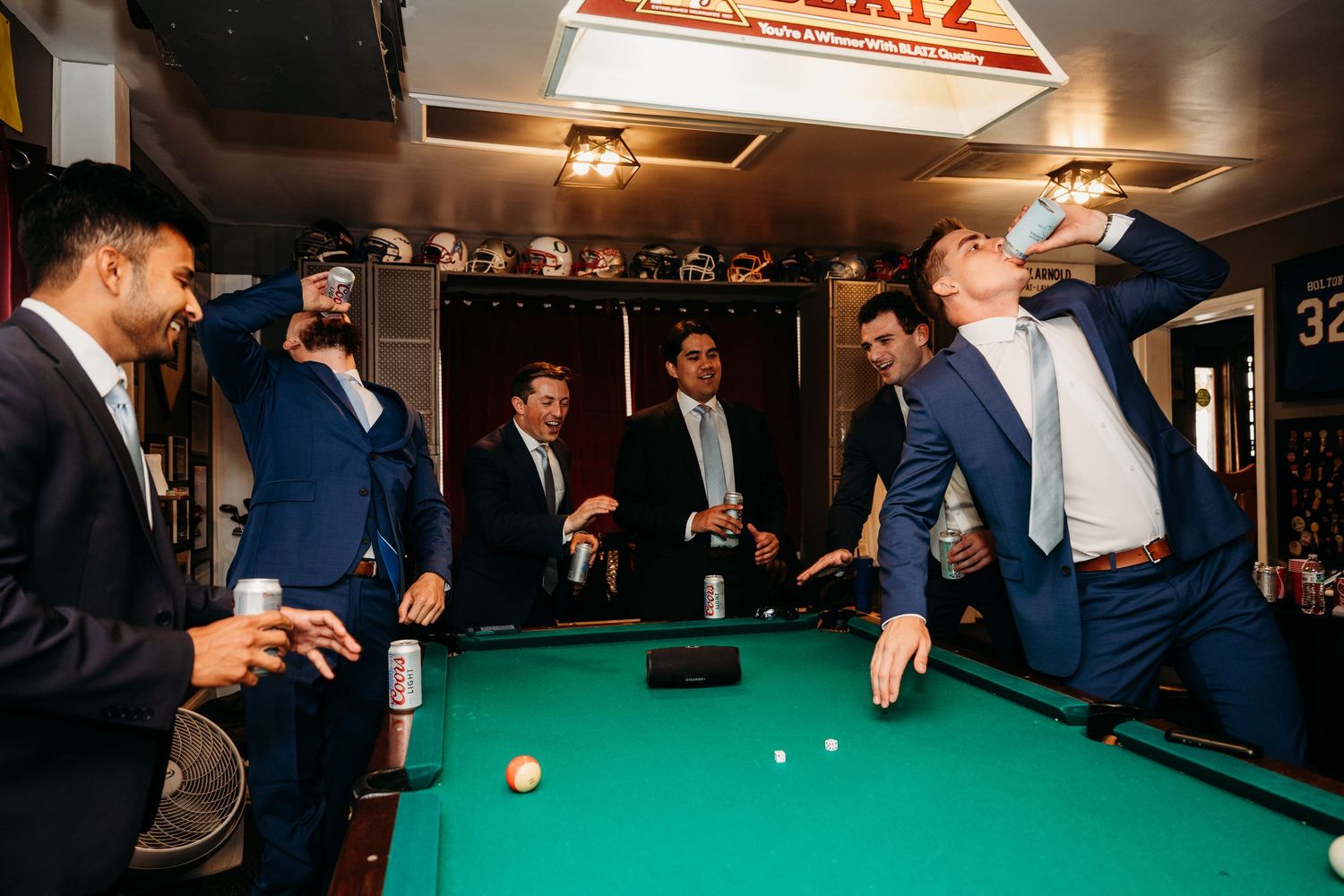 Groomsmen in navy suits laugh and play pool together at a wedding celebration party.