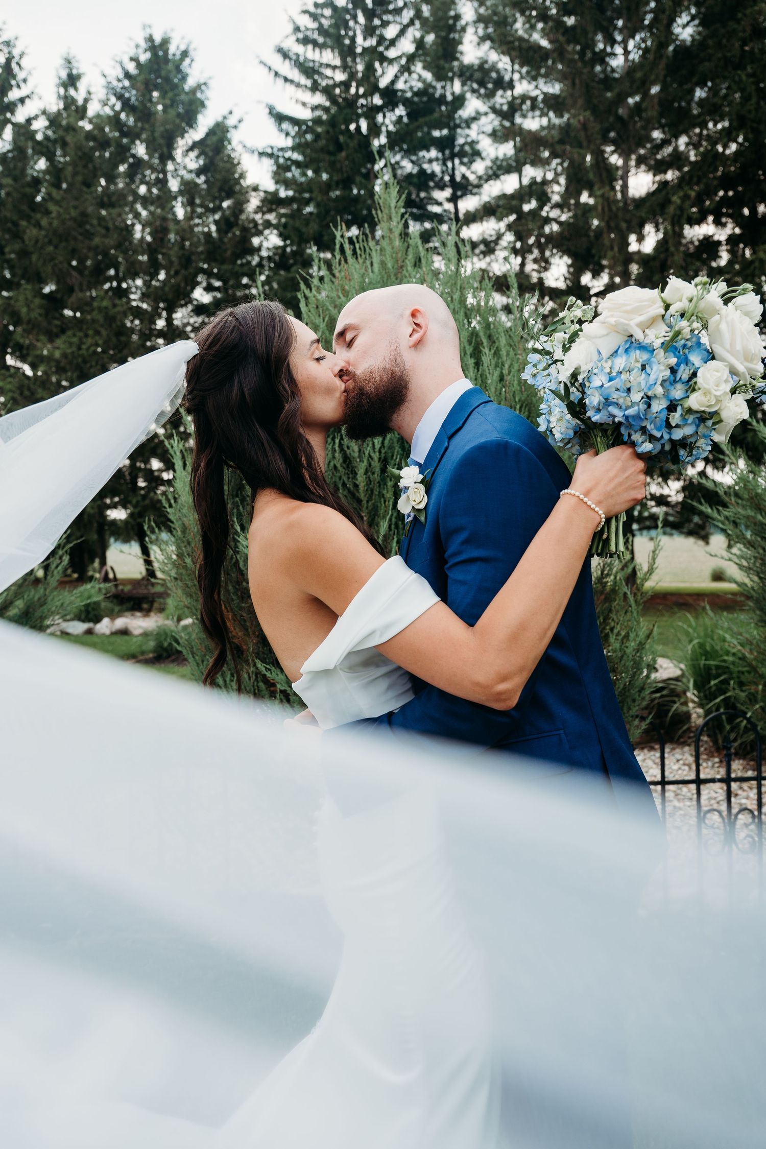 A wedding couple shares romantic kiss in garden setting with flowing veil and blue bouquet.