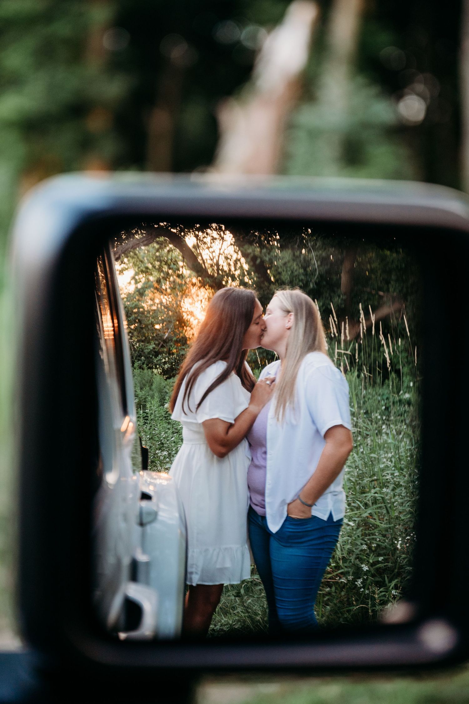 A romantic outdoor scene reflected in a car side mirror with sunlight filtering through trees in the background.