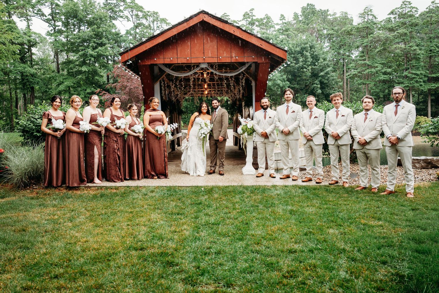 Wedding party poses before rustic wooden pavilion, bridesmaids in burgundy gowns and groomsmen in tan suits.