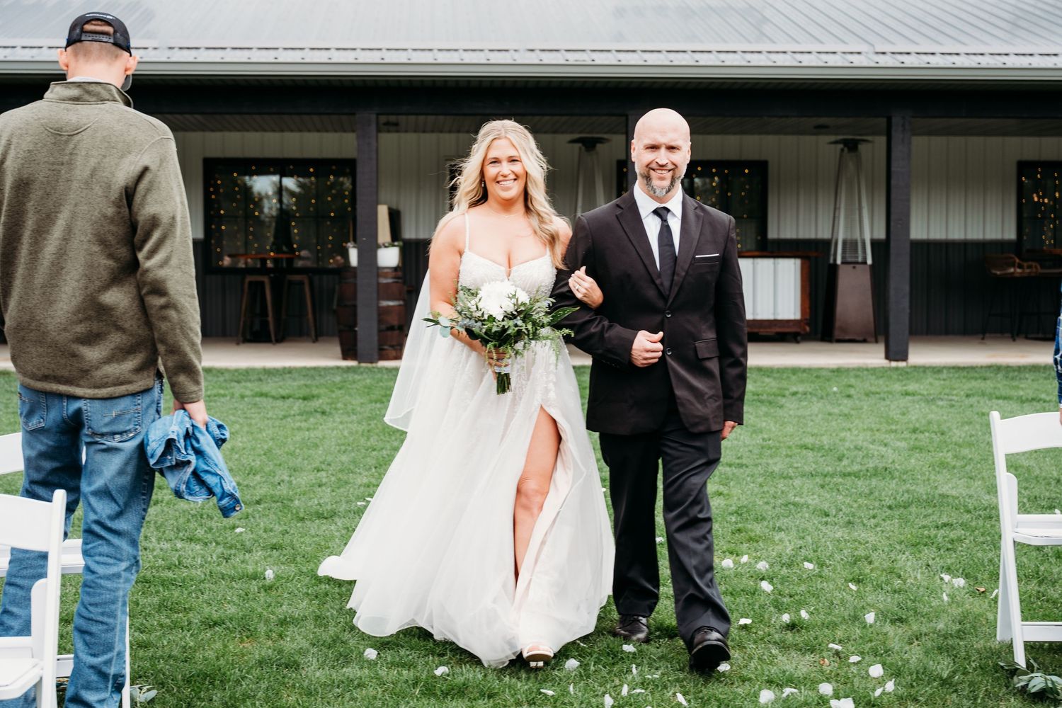 Bride in white gown walks down outdoor aisle with escort at rustic barn wedding venue.