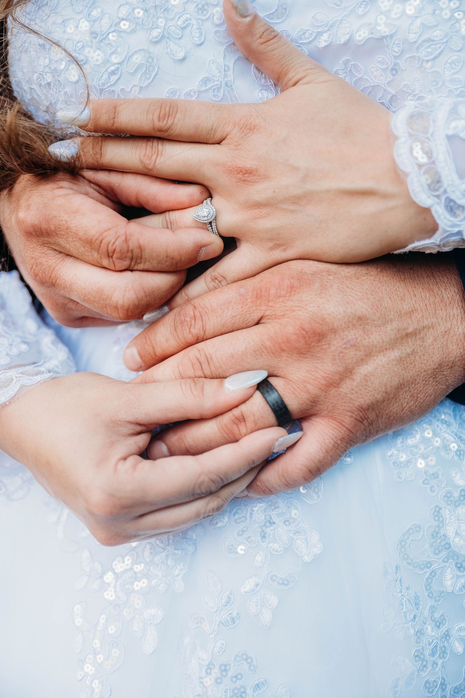 Close-up of newlywed couple hands showing diamond engagement ring and black wedding band on white lace bridal gown.