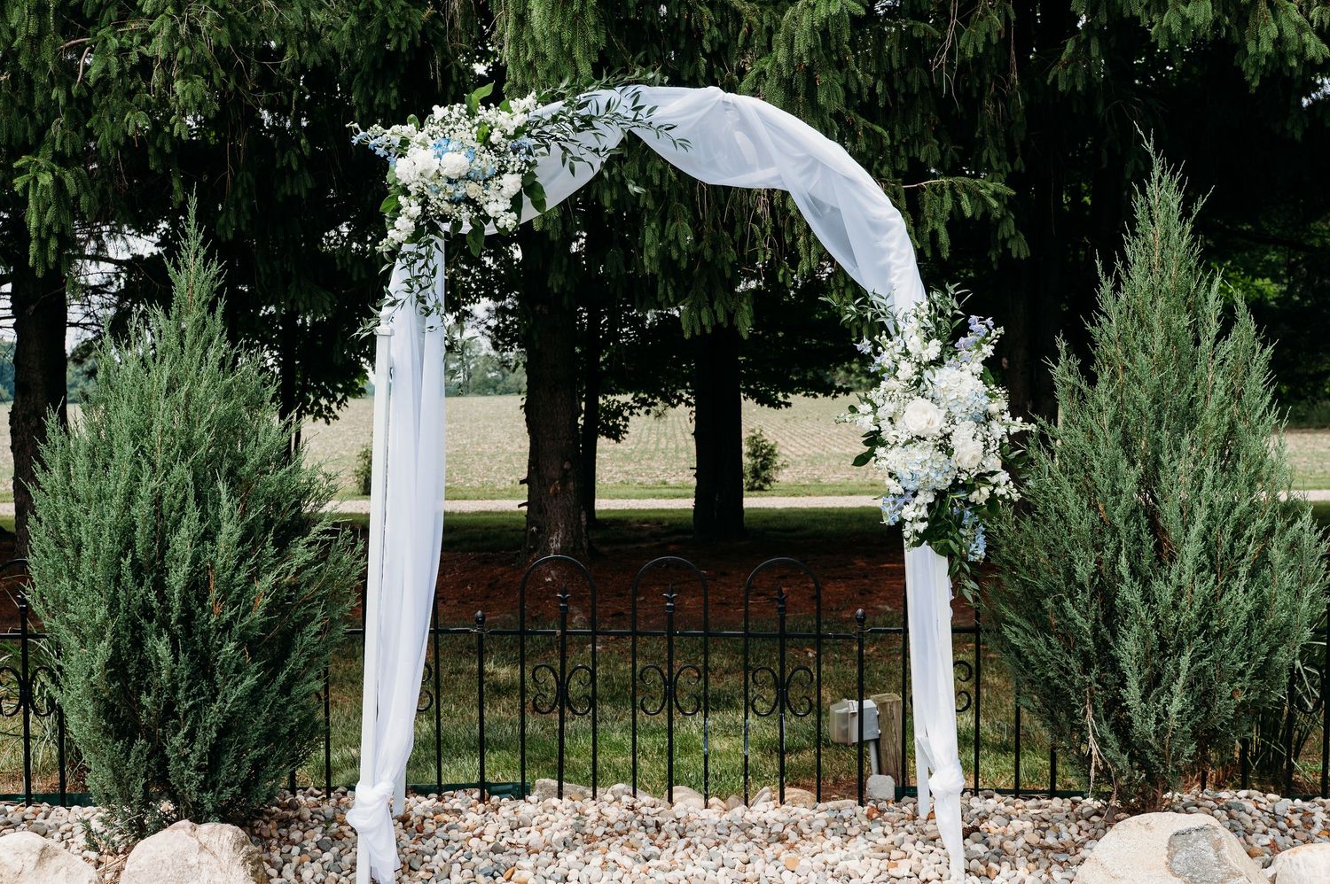 Romantic outdoor wedding arch adorned with white draping and floral arrangements surrounded by evergreen trees.