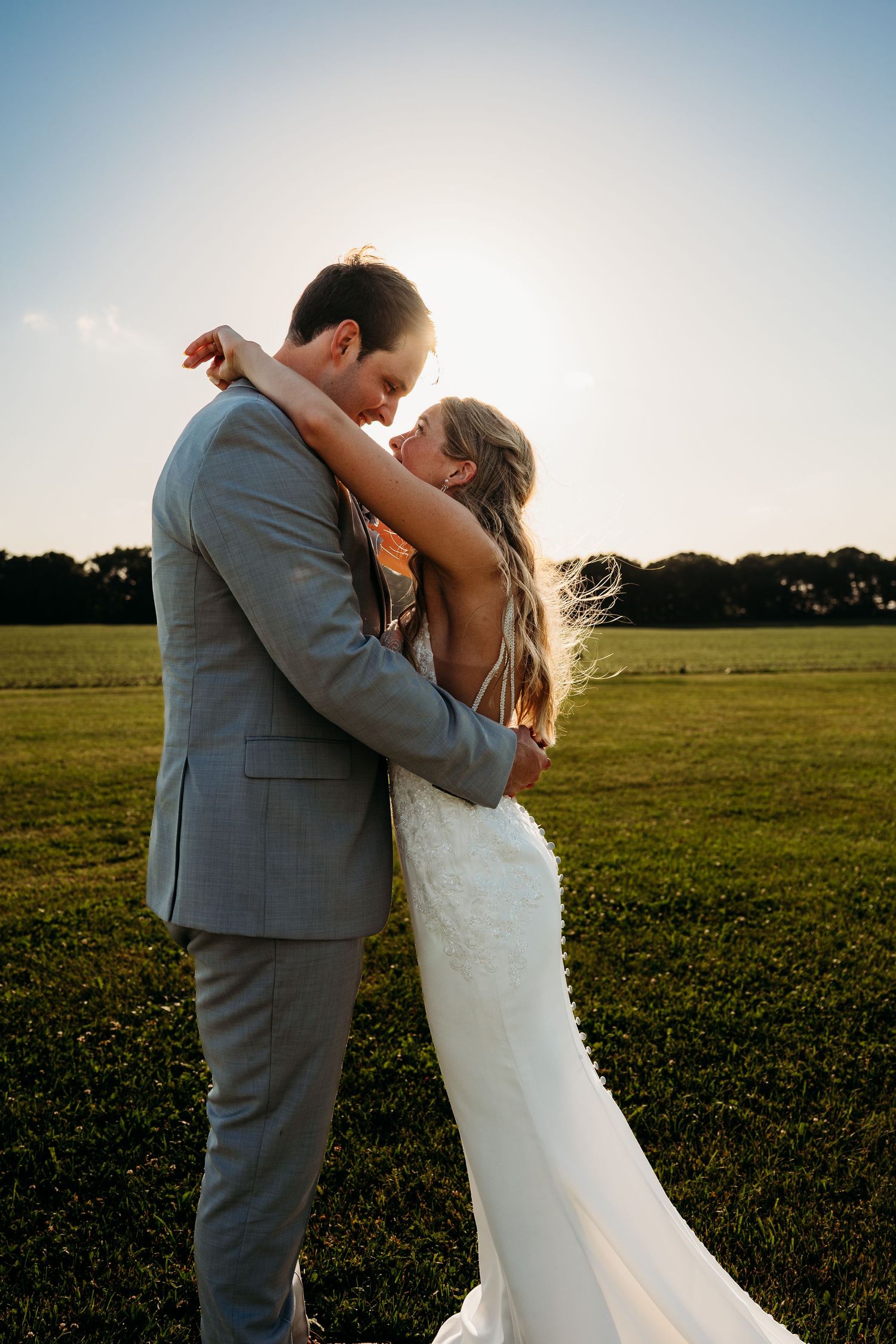 Bride and groom embracing at sunset in a green field, wearing a white gown and gray suit.