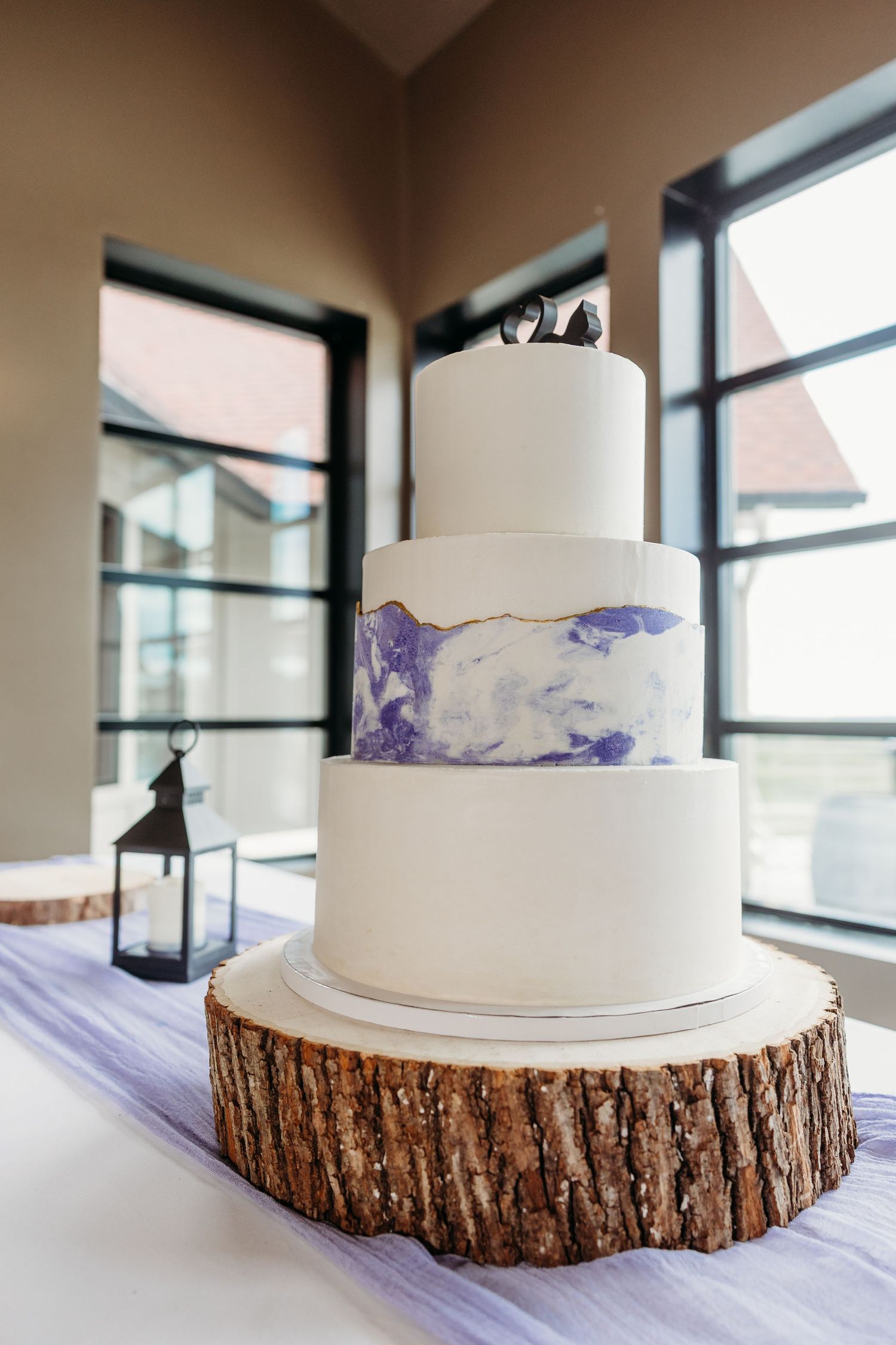 A white wedding cake with purple watercolor details sits elegantly on a rustic wood slice against a bright window backdrop.