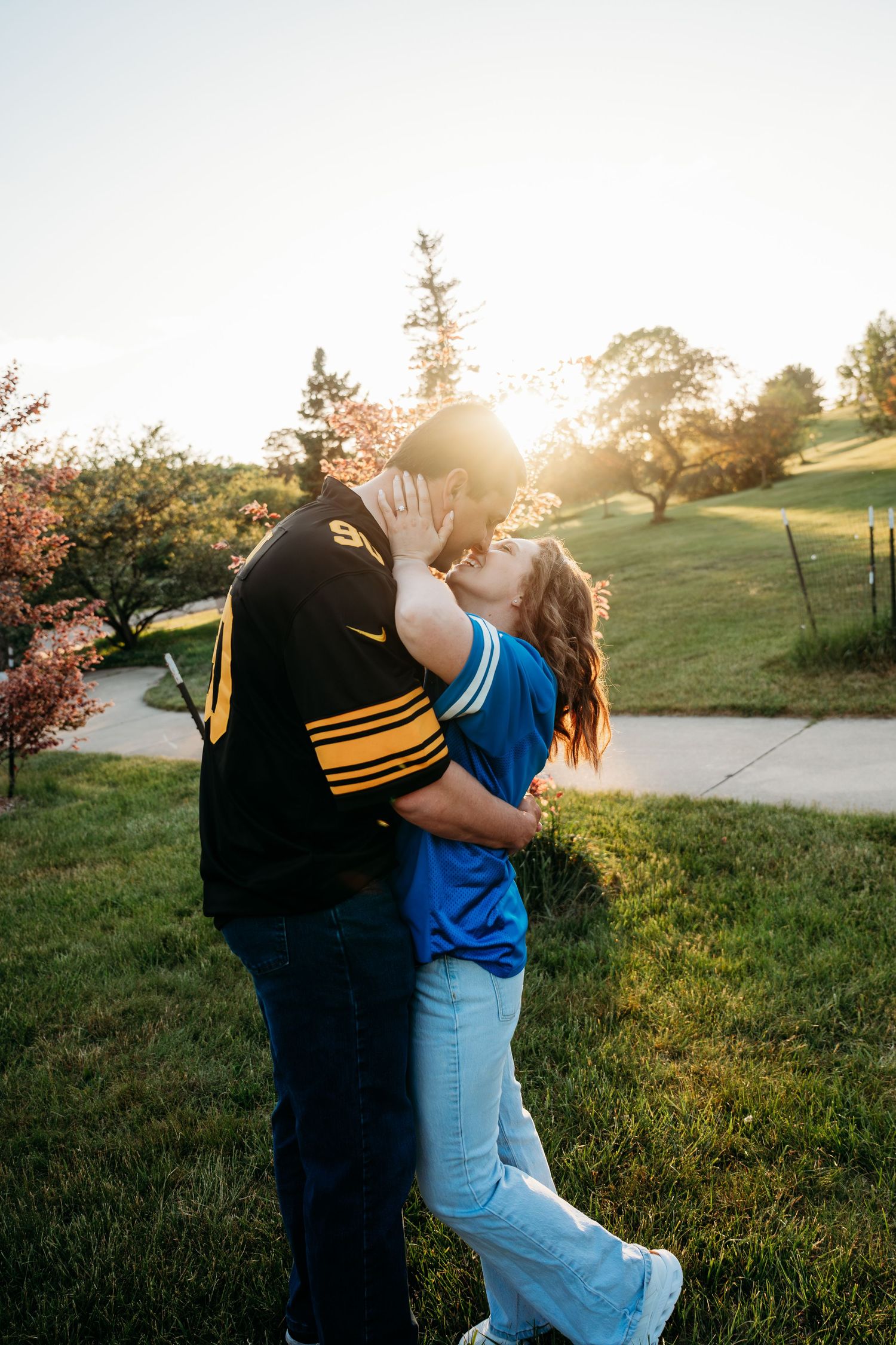 A couple shares a romantic kiss at golden hour on a lush green hillside, with warm sunlight glowing behind them.