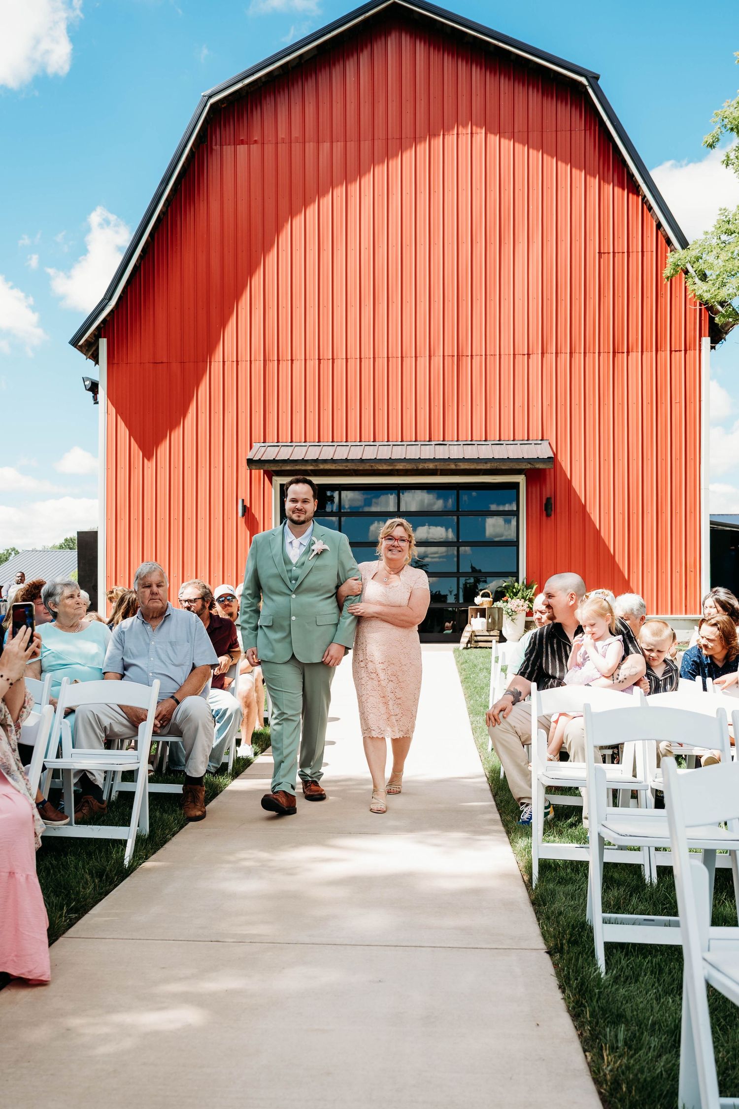 Groom in sage suit walks down outdoor aisle with mother before red barn wedding venue.