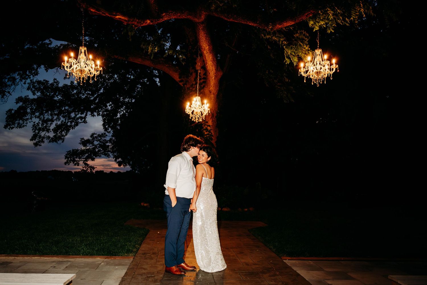 Couple sharing romantic kiss under glowing chandeliers hanging from large tree at outdoor wedding reception at dusk.