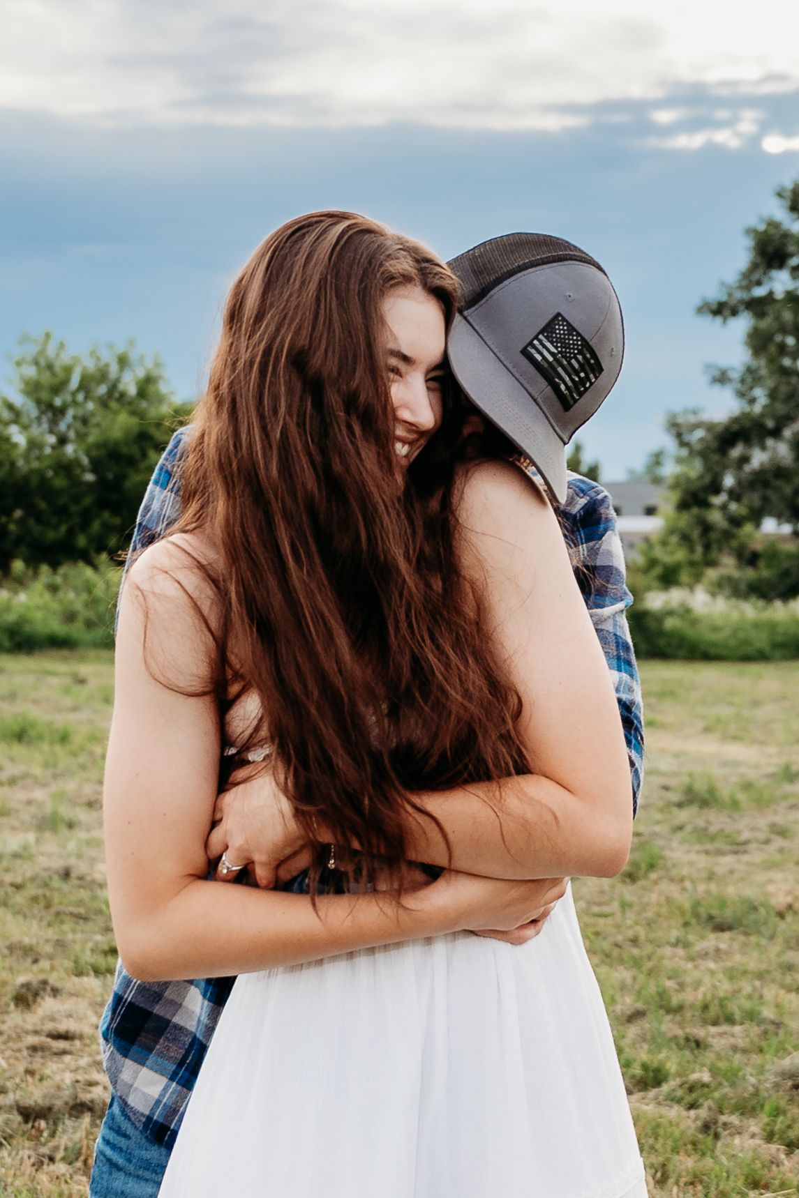 A couple embracing outdoors, woman in white dress with long hair smiling, man in plaid shirt and gray cap.