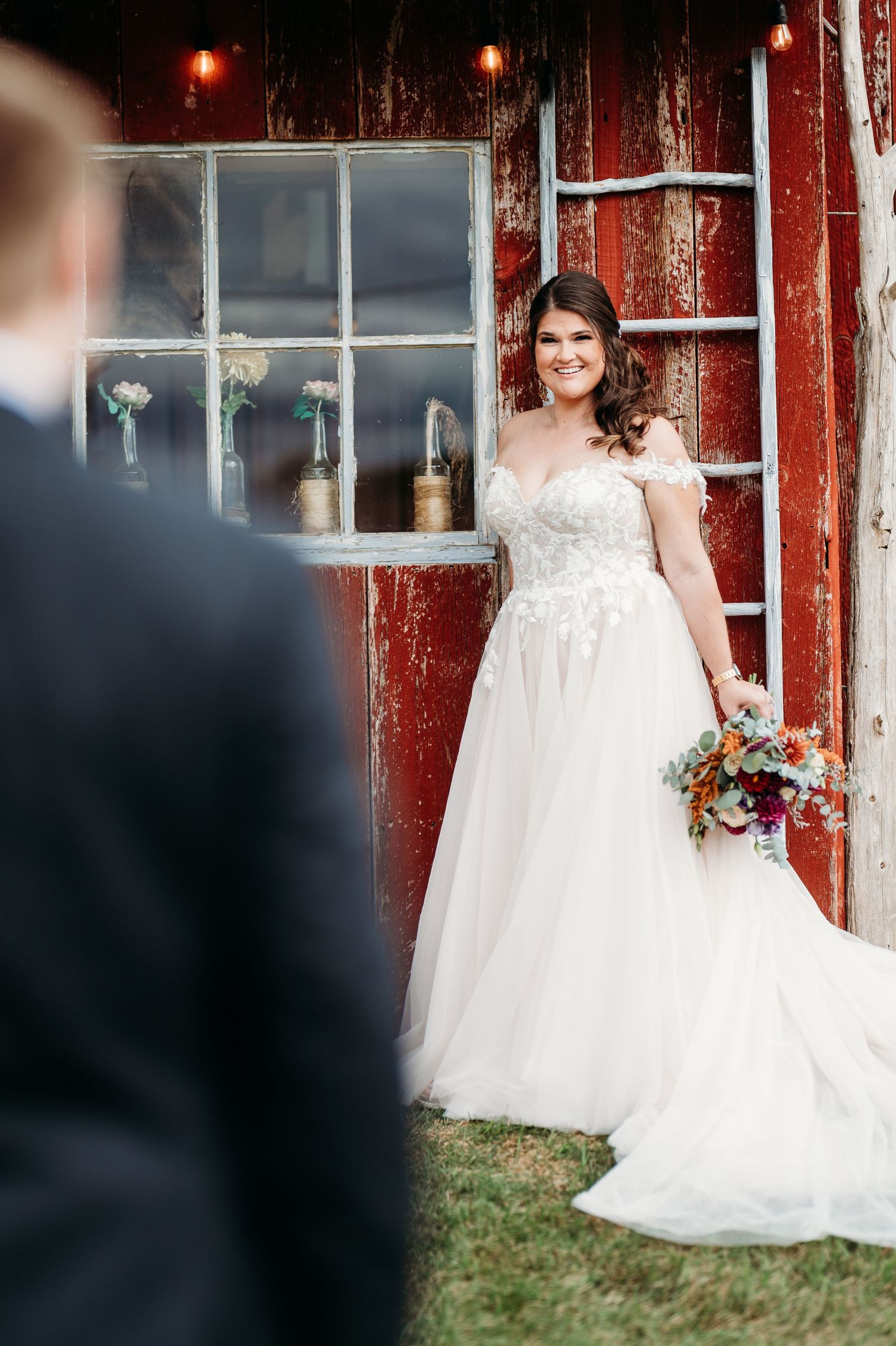 A bride in a flowing white gown poses against a rustic red barn with glowing lanterns.