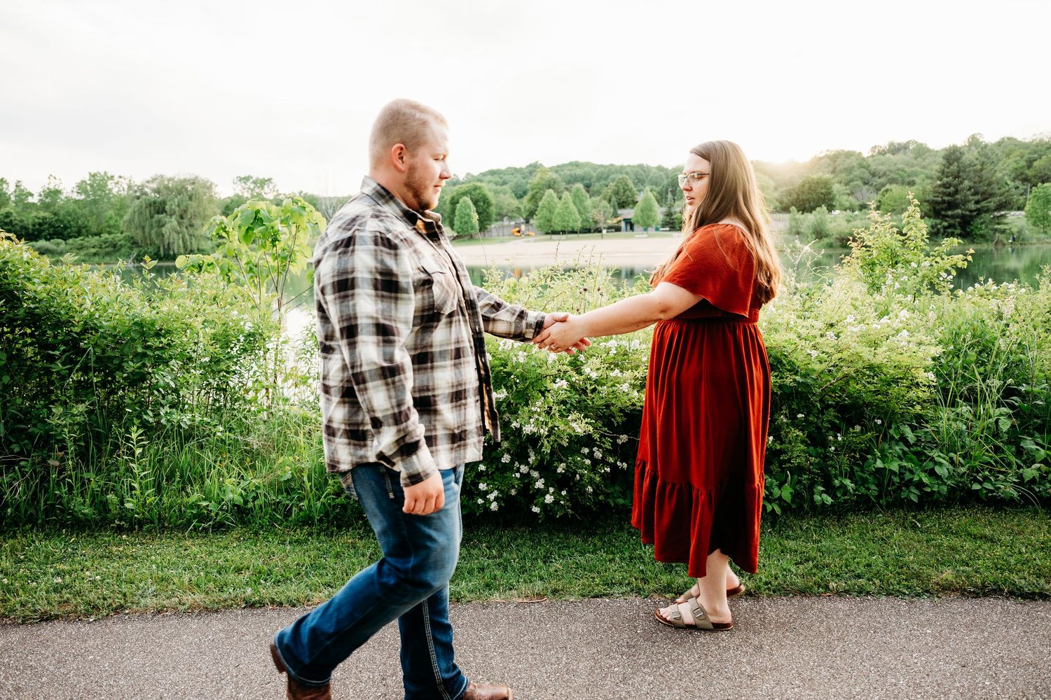 Couple holding hands walking on a path surrounded by lush greenery, man in plaid shirt and woman in red dress.