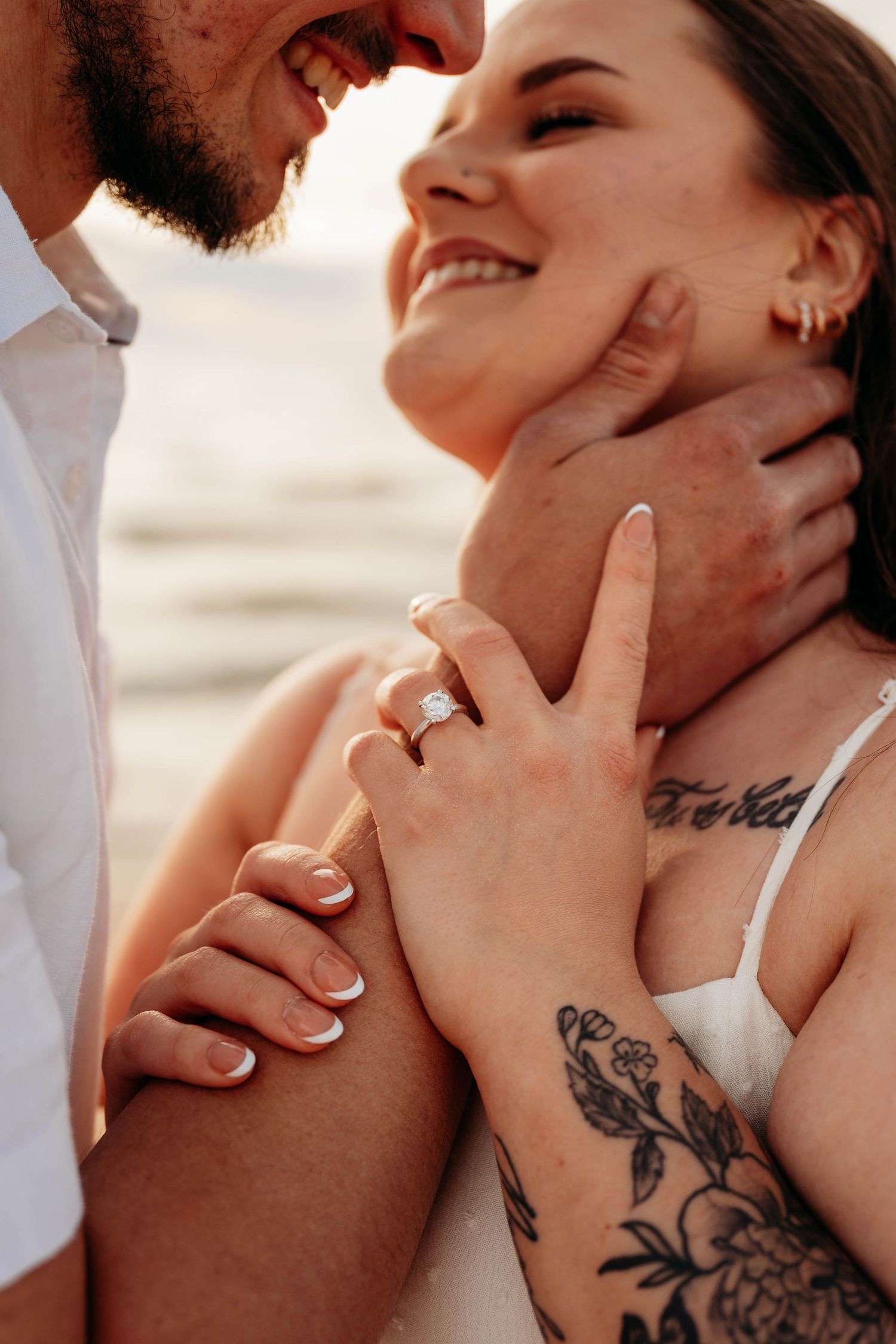 Smiling couple at beach showing engagement ring, woman with floral tattoo and French manicure embracing man in white shirt.