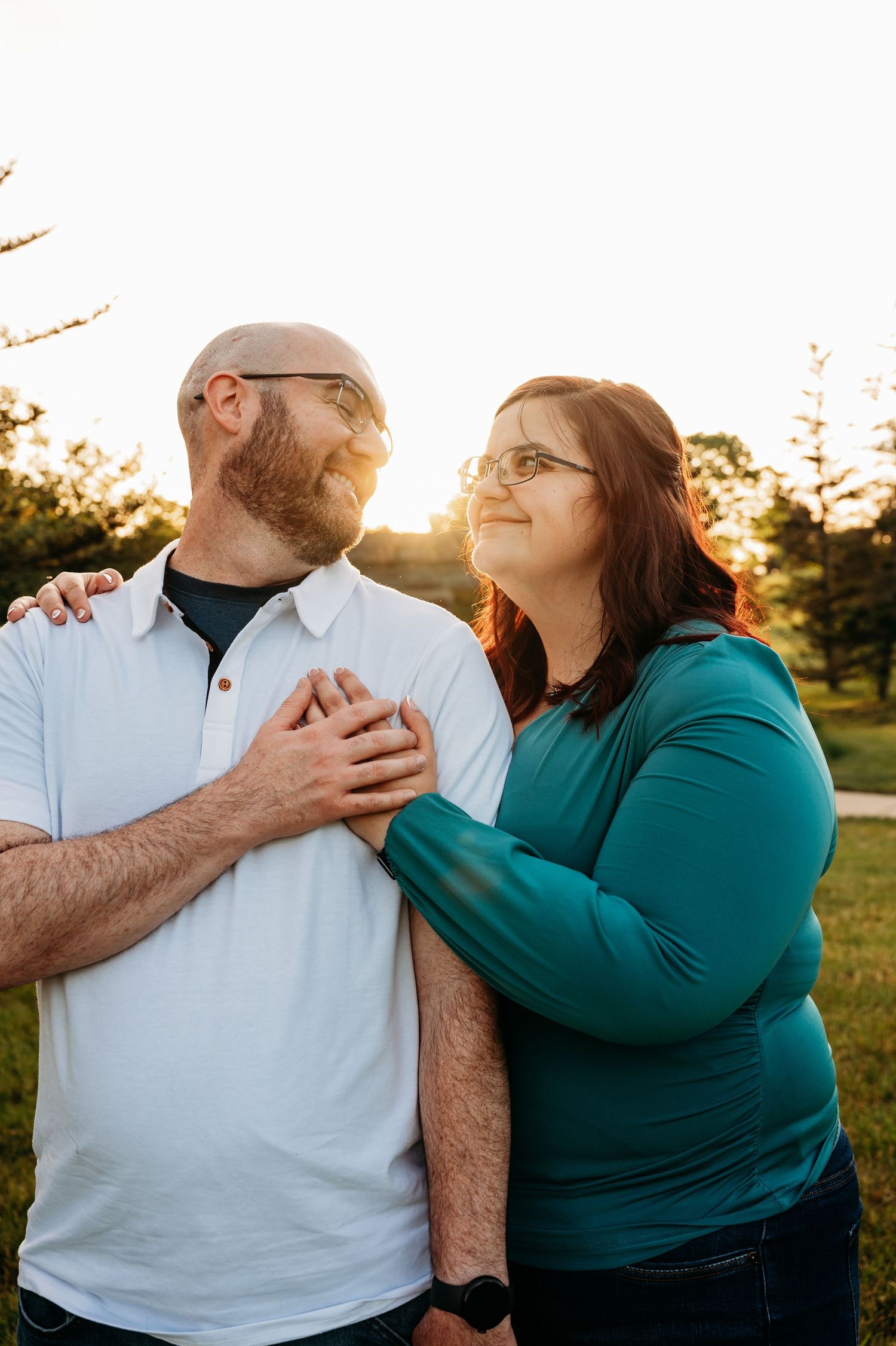 A couple stands together at golden hour, she in teal rests her hands on his chest as they share a tender moment.