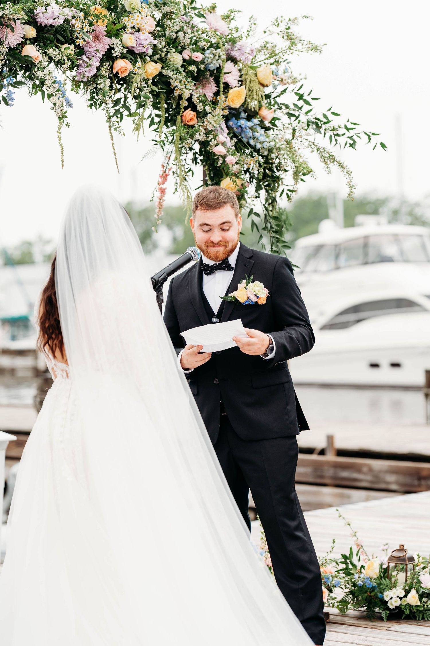 Groom in black tuxedo reads wedding vows to bride in white gown under floral arch at waterfront dock ceremony.