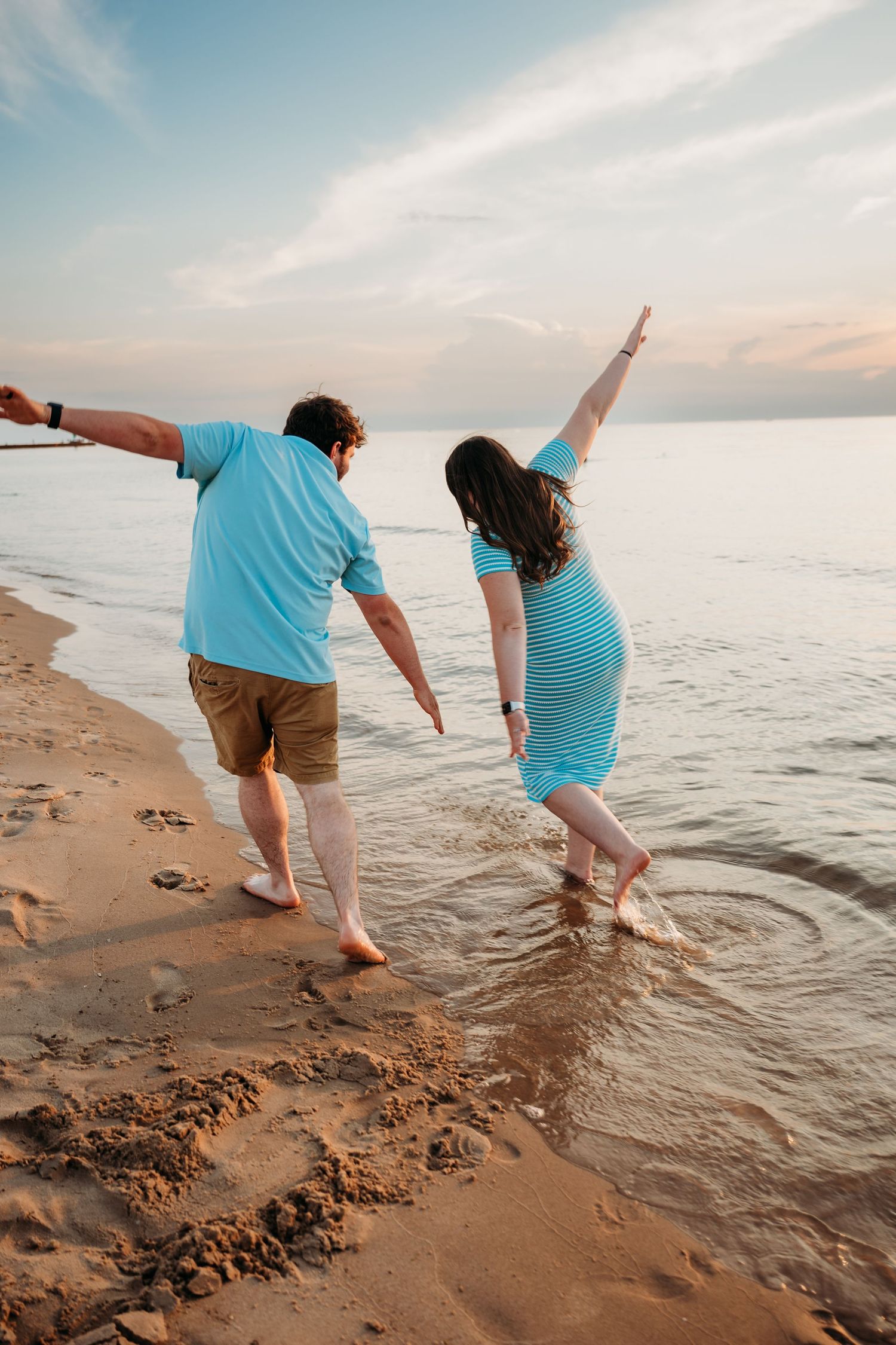 Two people wearing blue outfits playfully balance on one leg at the beach shore during sunset.