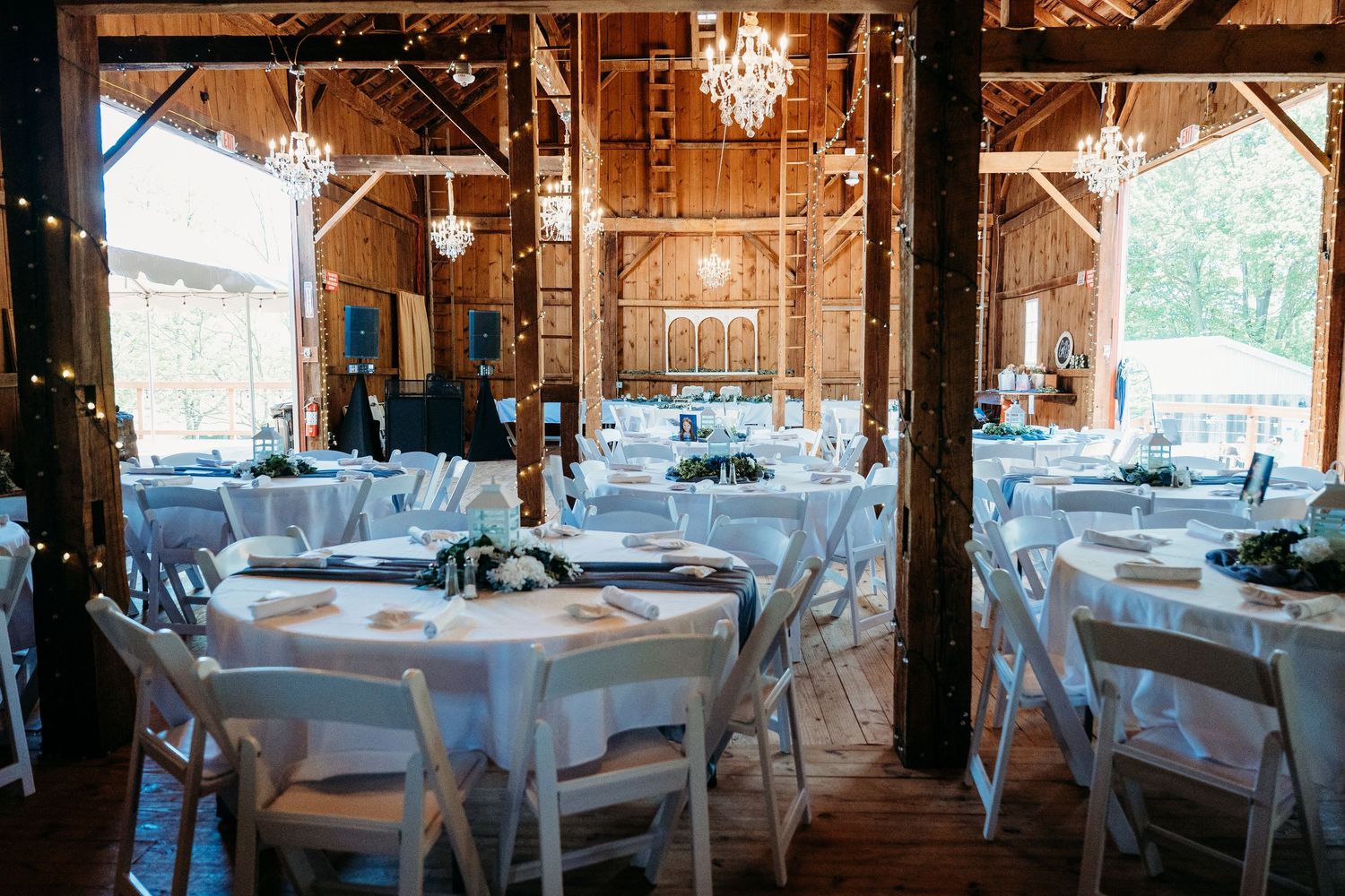 Rustic barn wedding reception venue decorated with elegant chandeliers and white tables set for dinner service.