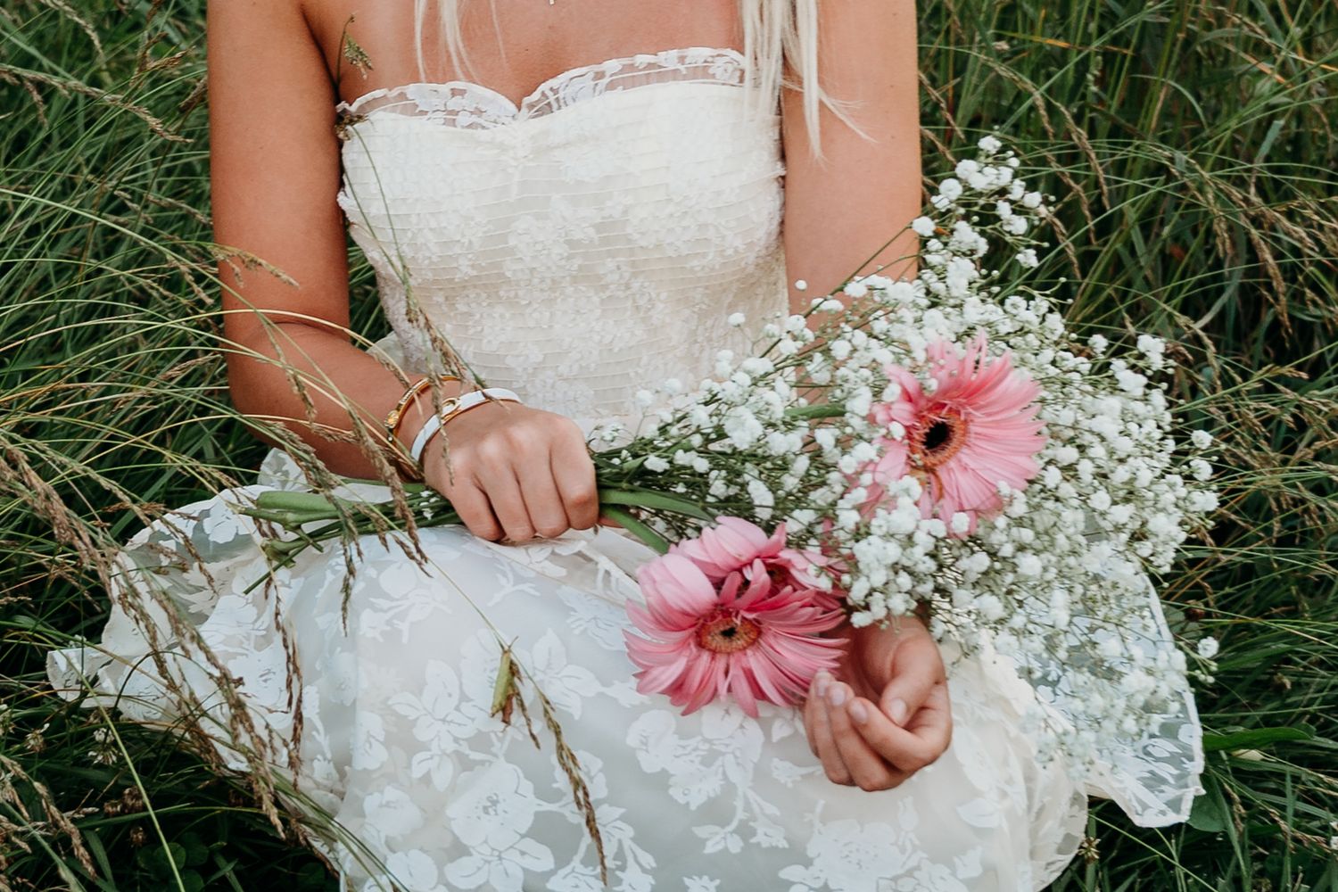 A wedding dress detail shows delicate white fabric and small flowers held against green grass in a natural setting.