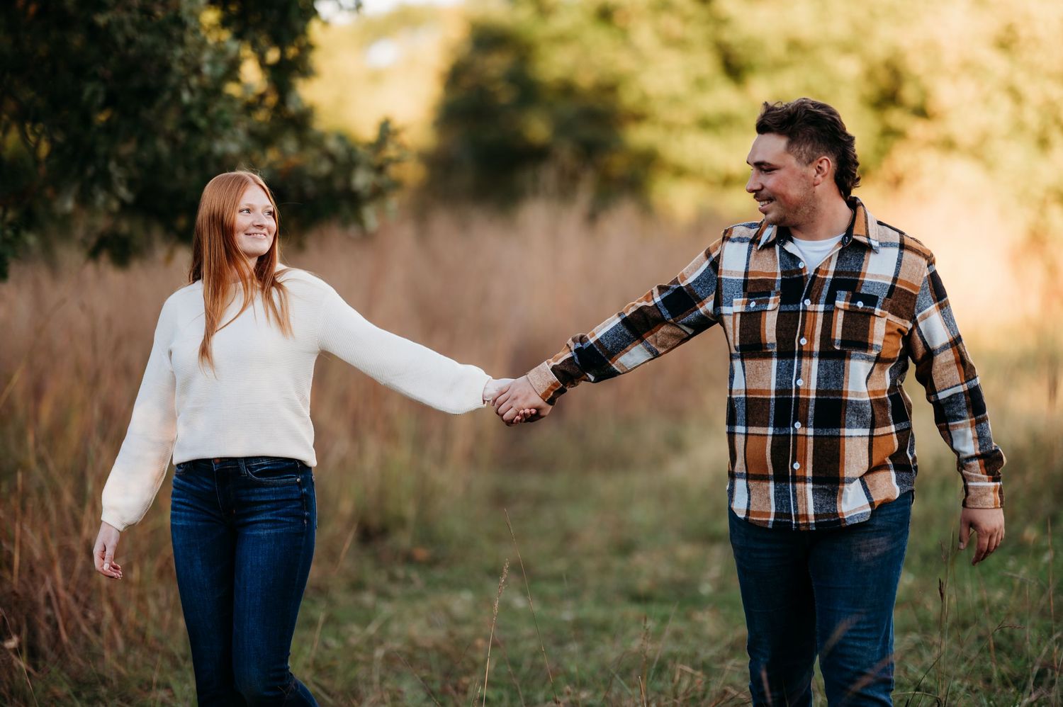 A couple holds hands and walks through a golden sunlit field during a romantic autumn photoshoot.