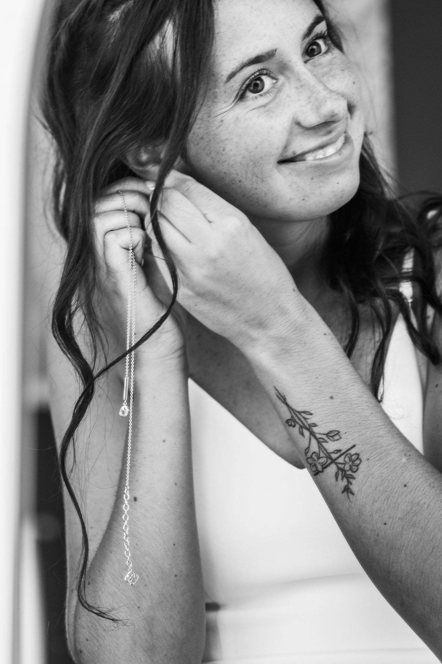 Black and white portrait showing a natural smile and delicate floral tattoo on forearm.