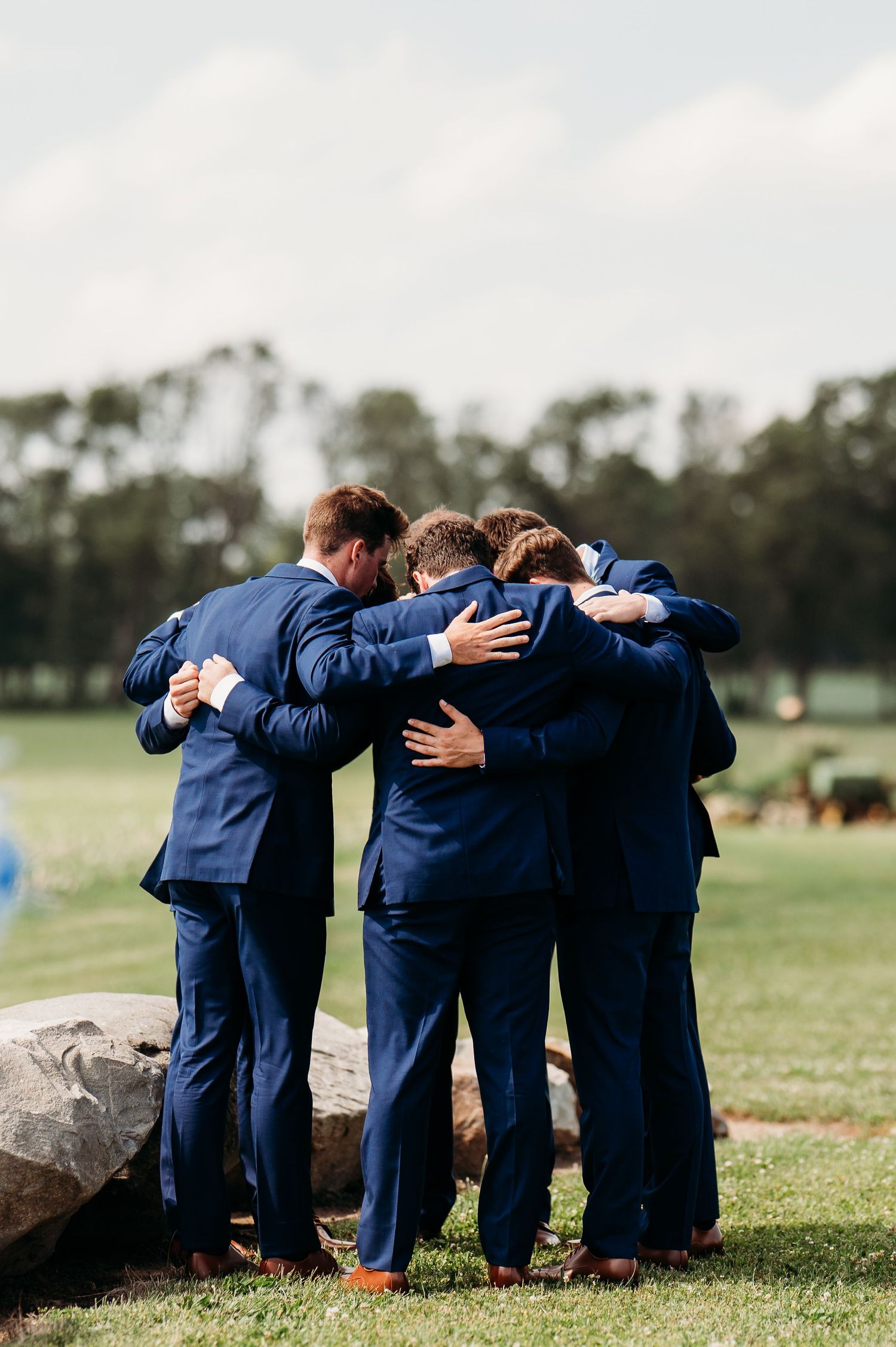 A group of groomsmen in navy blue suits share an emotional embrace in a circle on grassy field.