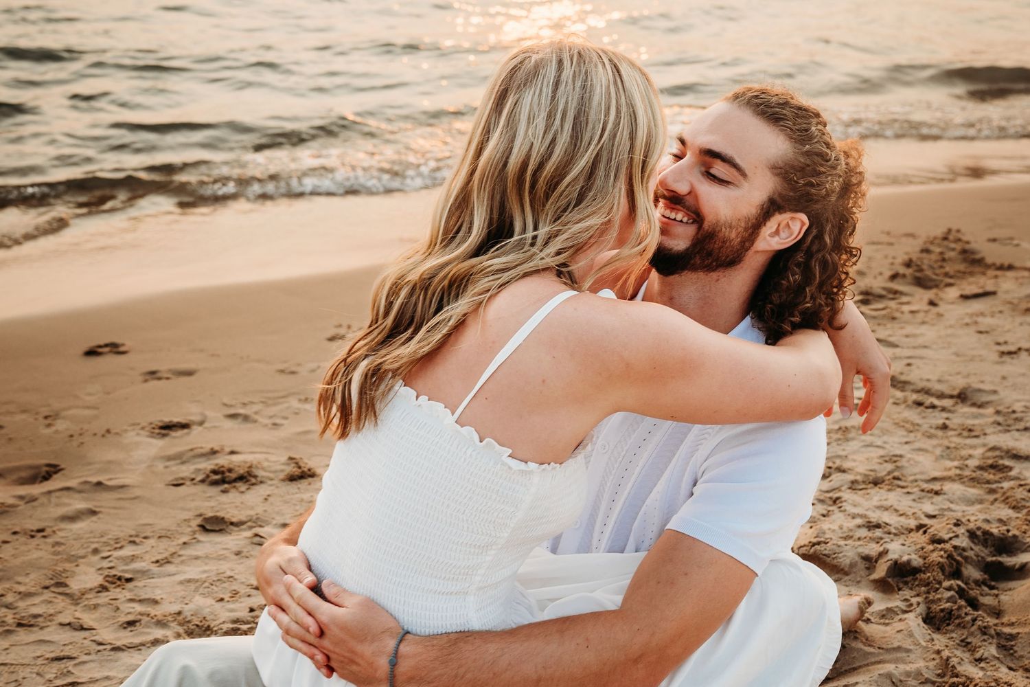 A couple in white embraces on a sandy beach at golden sunset, sharing a romantic and joyful moment.