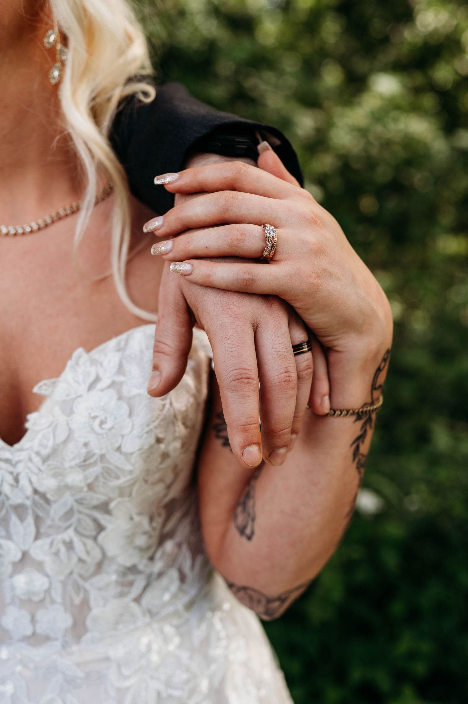 Close-up of hands wearing wedding rings and engagement ring, with lace dress detail and small wrist tattoo visible.