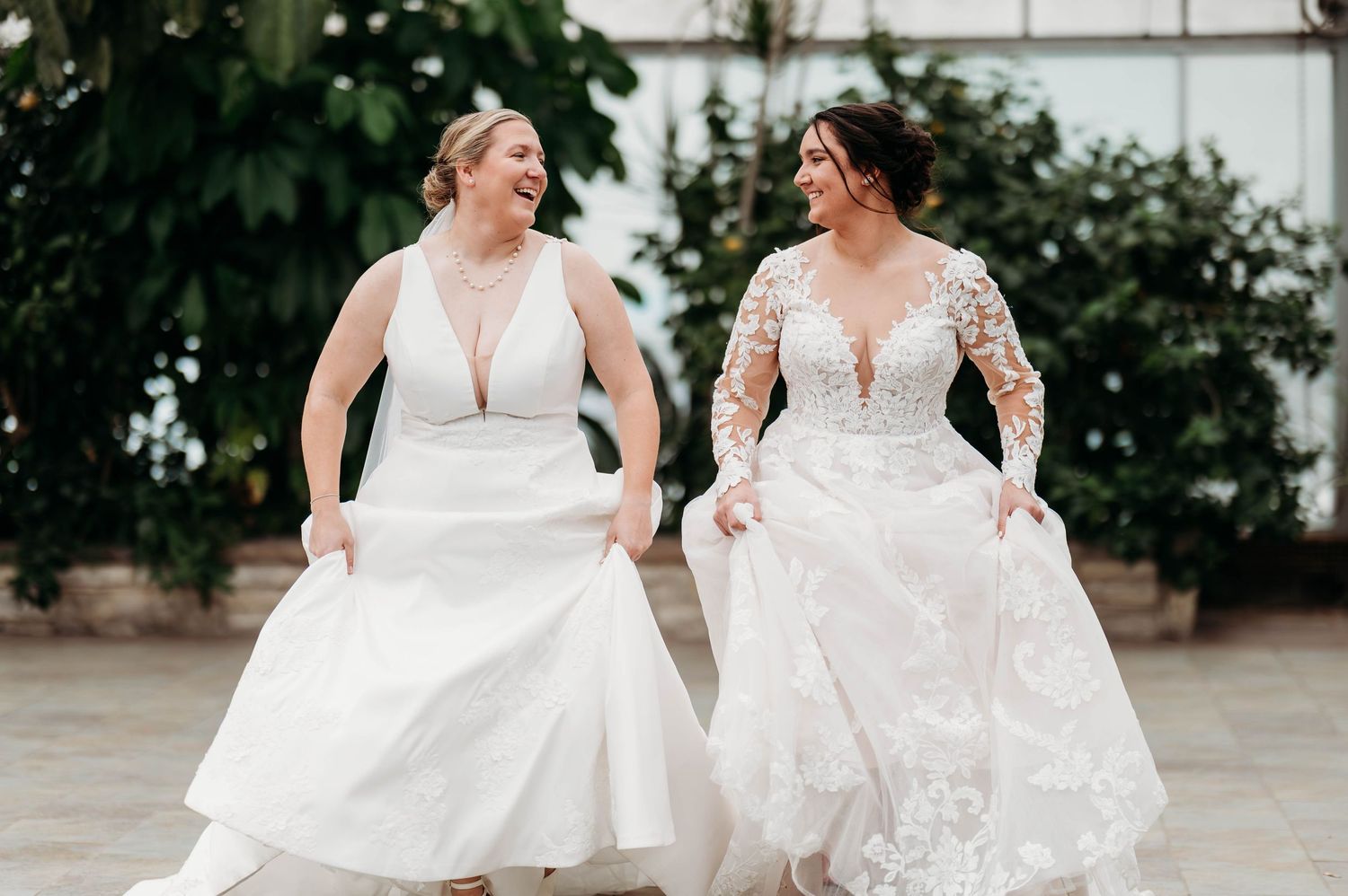 Two brides in flowing white dresses laugh together while twirling their gowns during a wedding photoshoot.