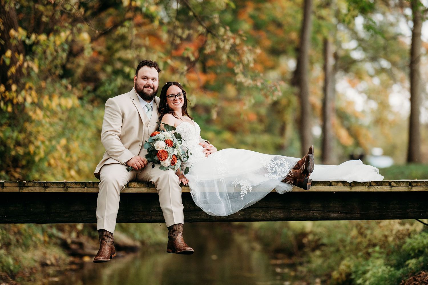 A couple in wedding attire sits together on a wooden bridge surrounded by autumn foliage in a romantic outdoor setting.