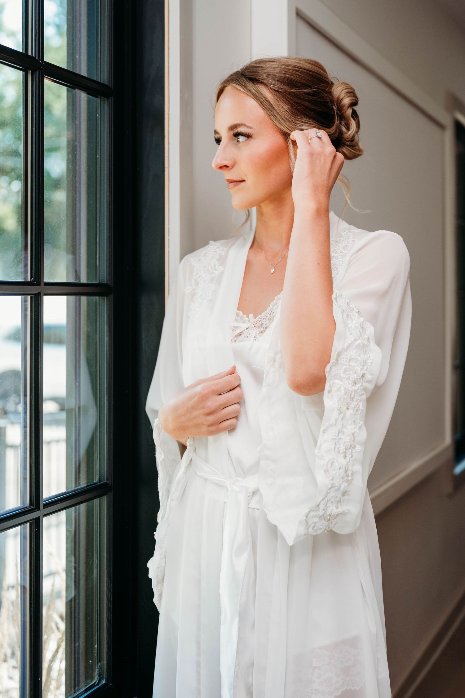 Bride in white lace robe adjusting earring by window, elegant updo hairstyle, soft natural light.