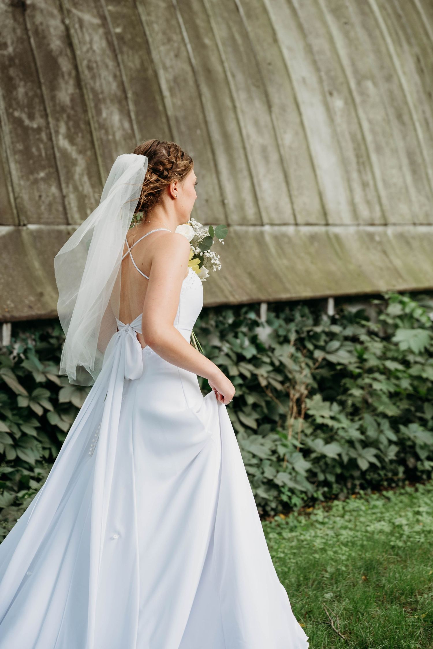 Bride in white satin gown and veil holds bouquet, viewed from behind in lush garden setting.