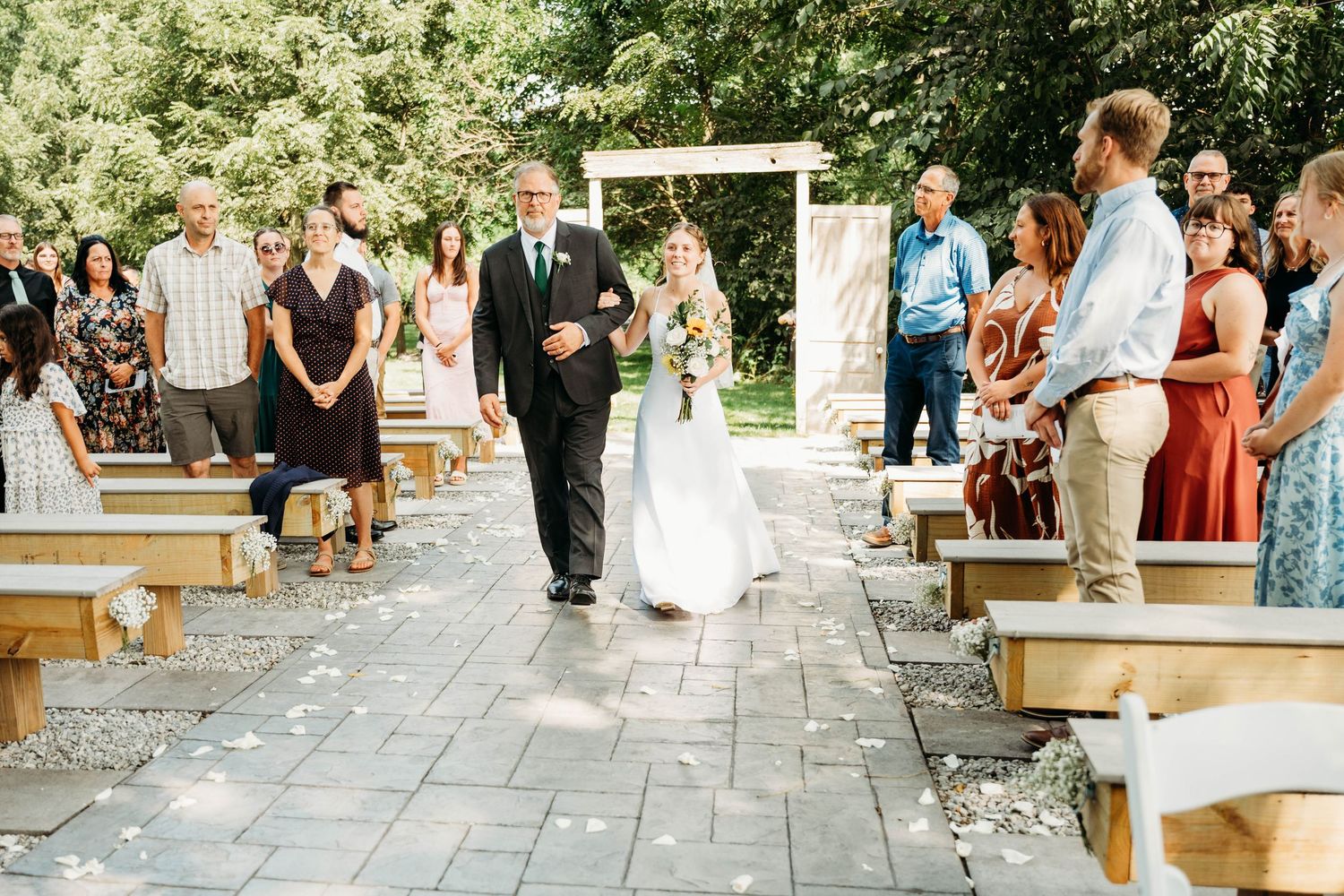 Bride walks down the aisle with her father at an outdoor wedding ceremony while guests watch.