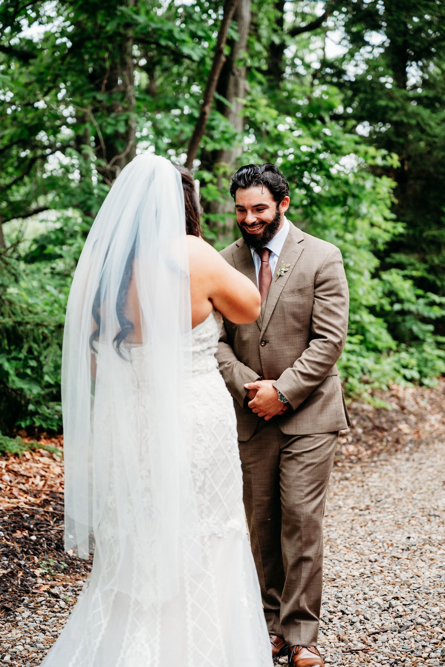Groom in tan suit smiles at bride in lace wedding dress and veil during outdoor first look ceremony.