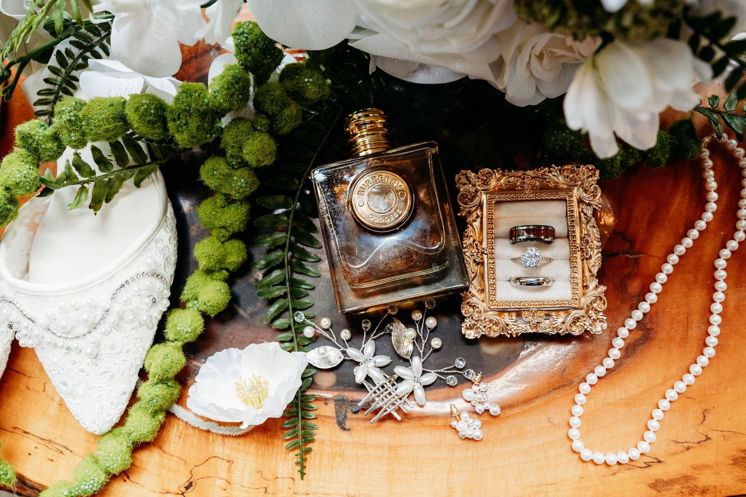 Elegant wedding flat lay with perfume bottle, gold ring frame, pearl necklace, floral hair pins, and white flowers on wood.