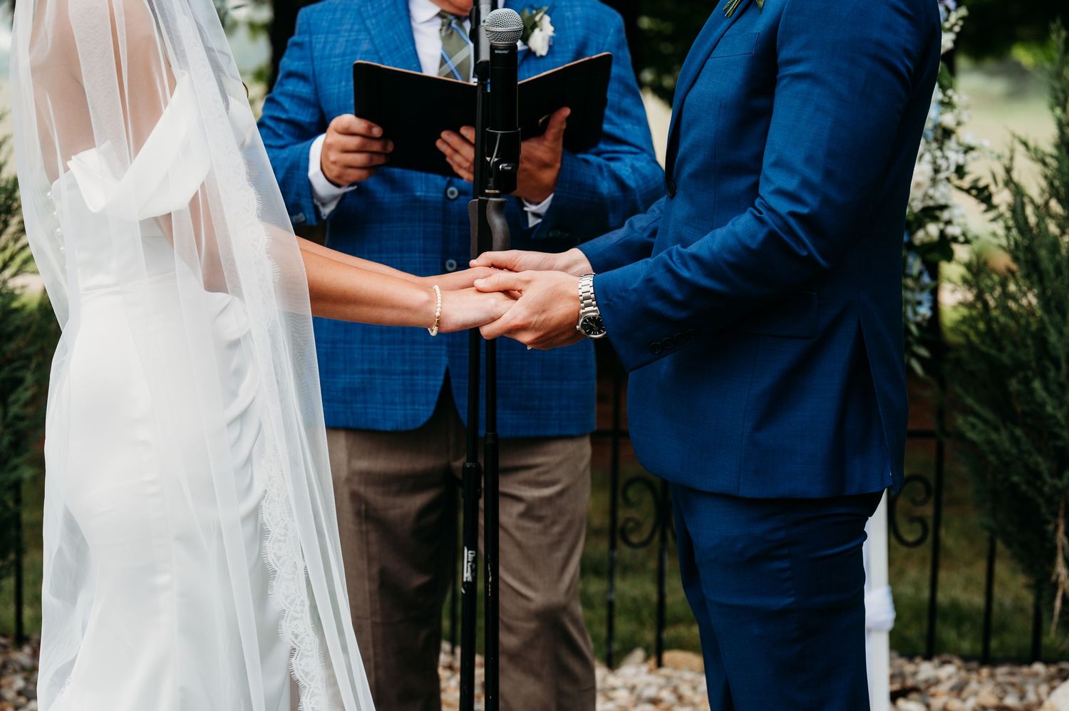 A couple holds hands during an outdoor wedding ceremony with an officiant in a blue suit holding a book.