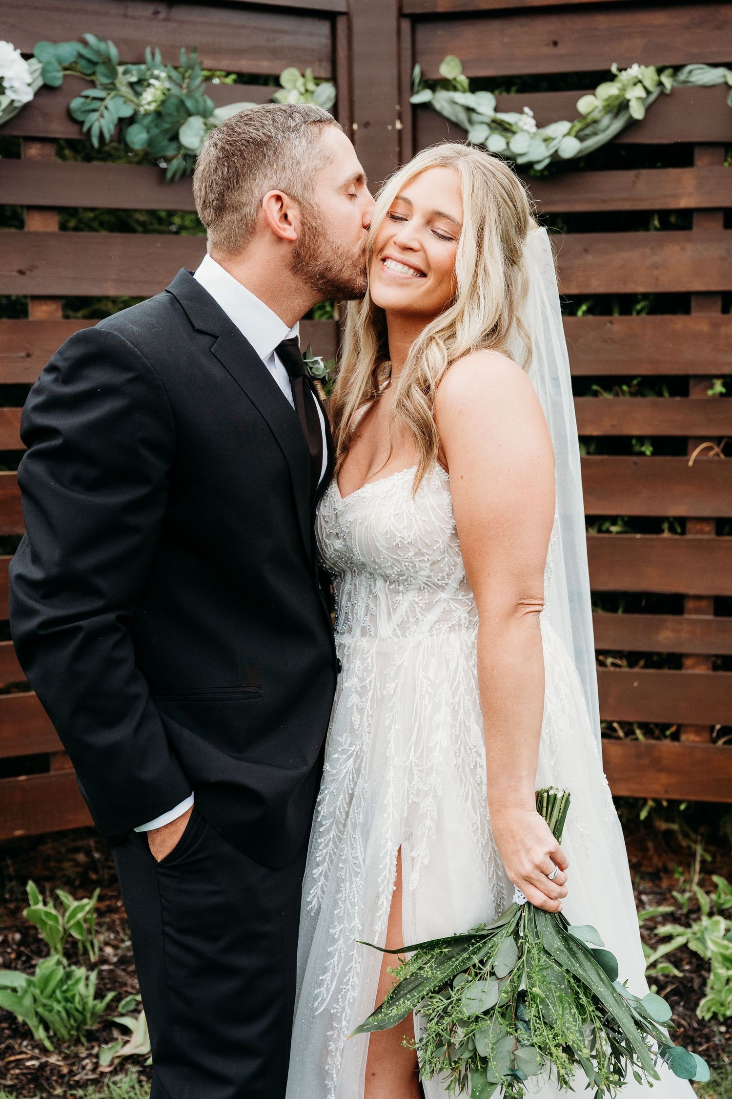 Groom in black suit kisses bride in lace wedding dress holding greenery bouquet before wooden fence backdrop.