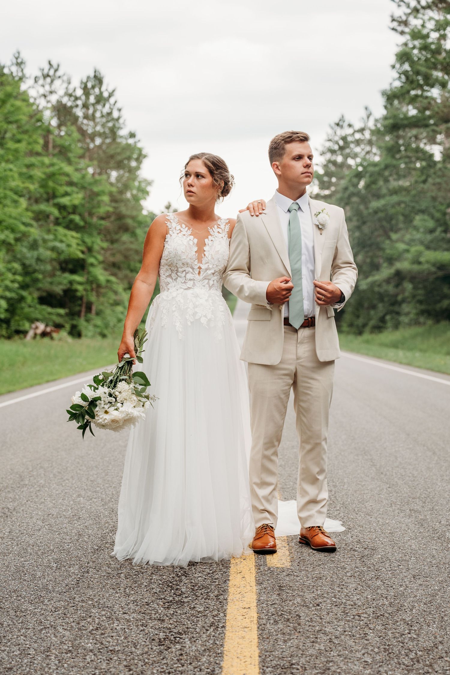 Bride and groom standing on a road surrounded by trees, wearing white gown and beige suit with sage tie.