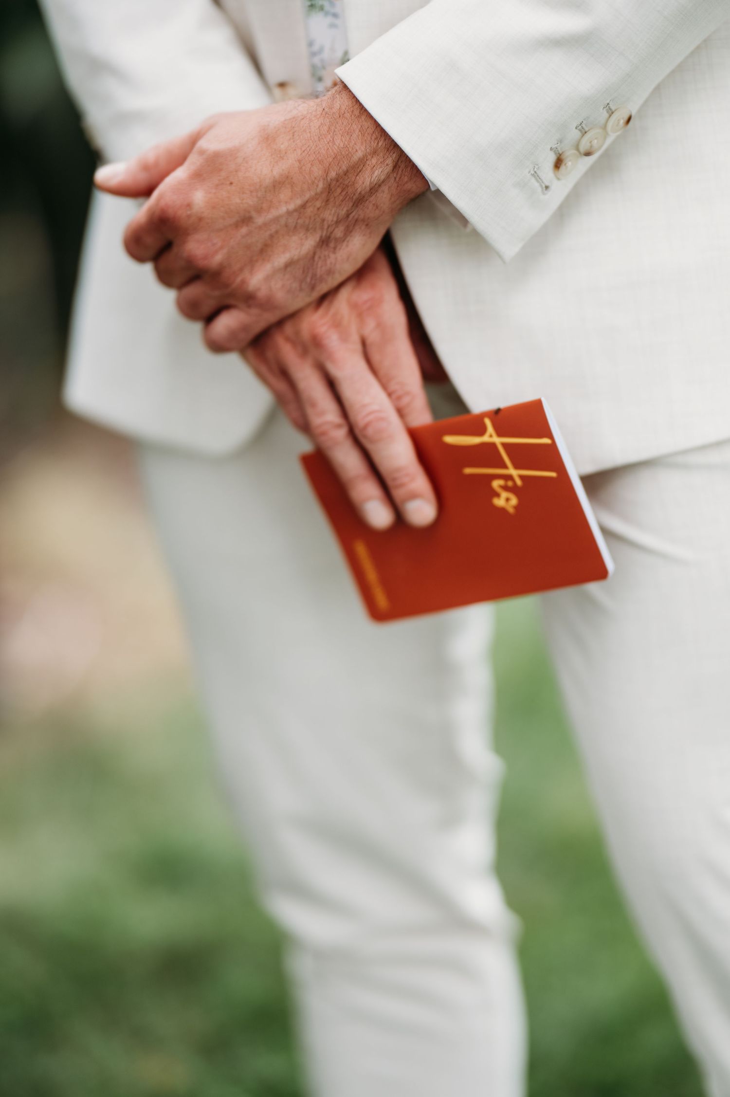 Groom in white suit holding a red vow book with gold cross detail during outdoor wedding ceremony.