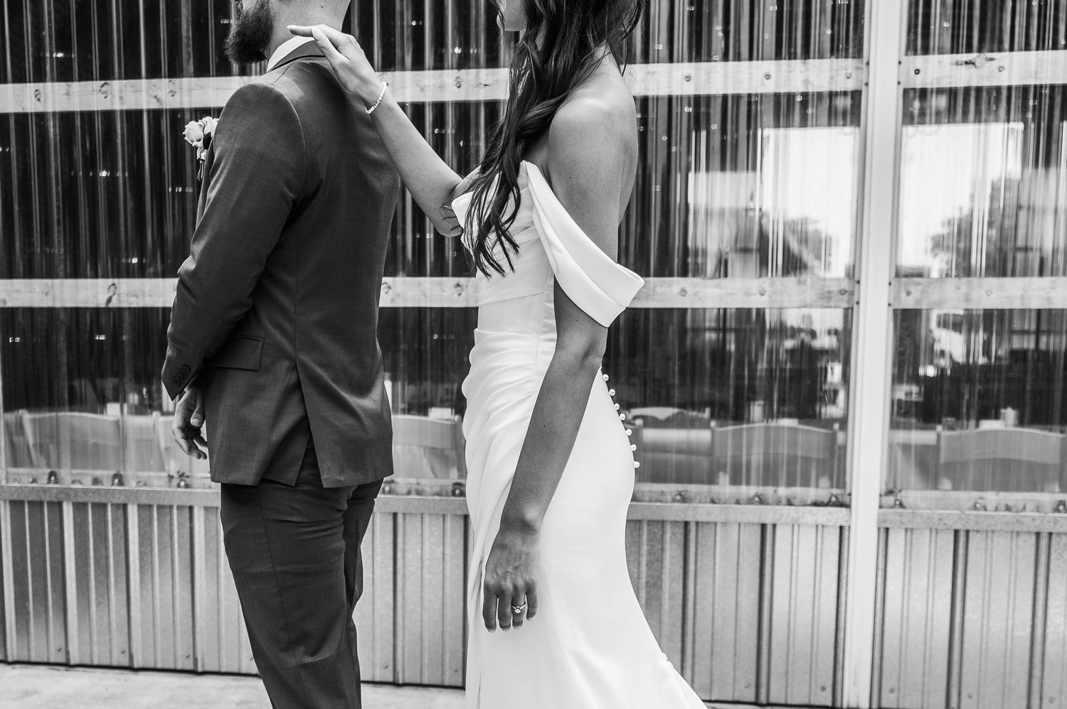 A bride in an elegant off-shoulder white gown stands with a groom in a dark suit in a black and white wedding photo.