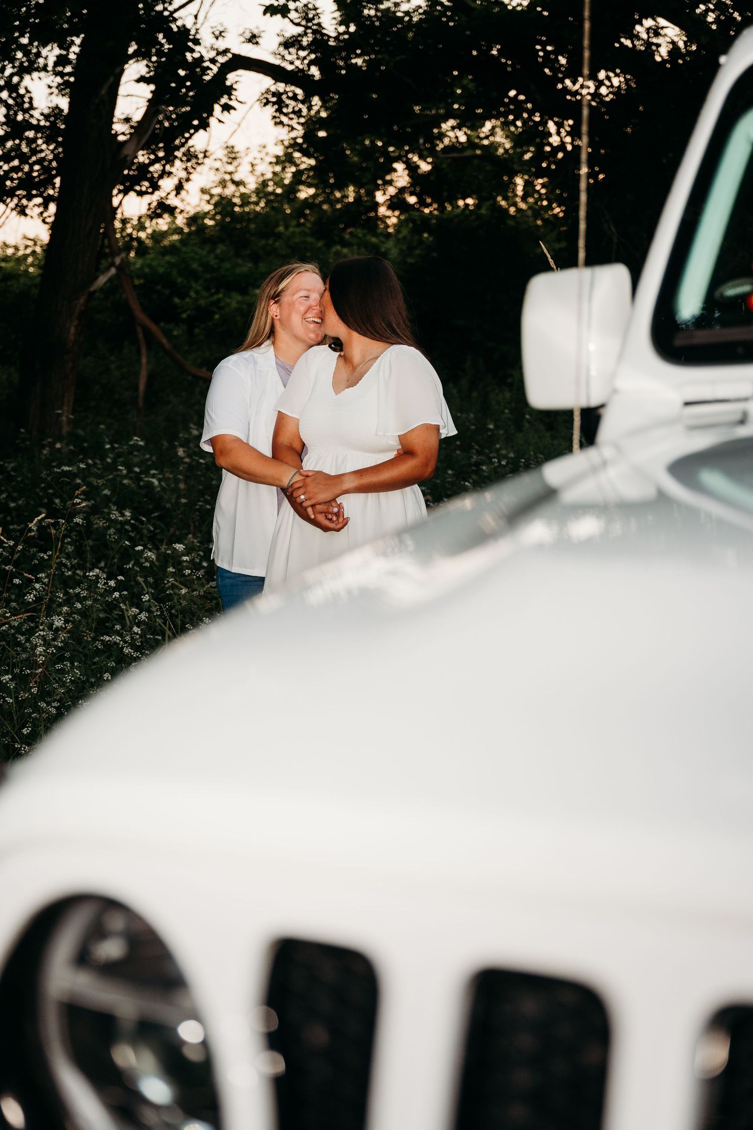 People in white clothing stand next to white vehicles parked near trees at dusk creating a monochromatic scene.
