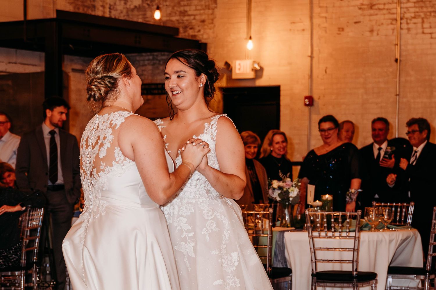 Two brides share their first dance together at a rustic brick venue illuminated by warm lighting during their wedding reception.