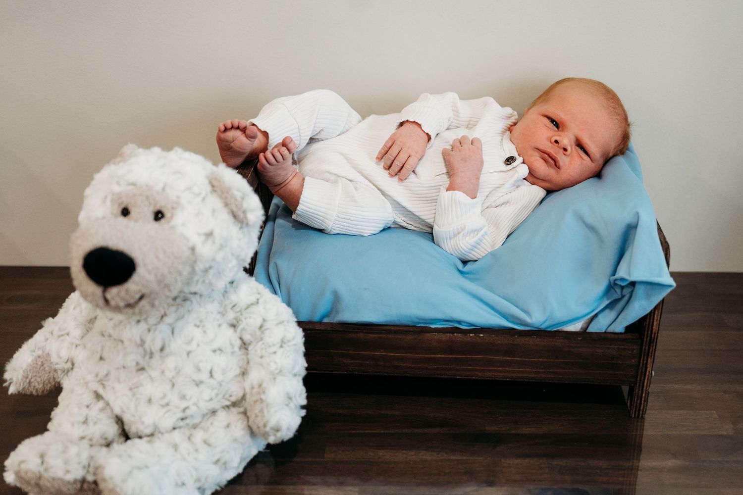 A sleeping baby in white clothing rests on a blue cushion next to a plush white teddy bear on a wooden surface.