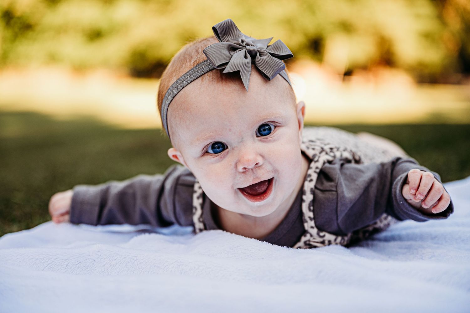Baby wearing gray outfit and bow headband smiles while lying on blanket outdoors.