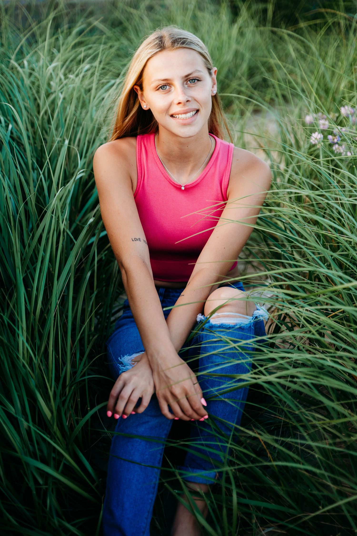 A person in pink athletic tank top and blue leggings sits among tall green grass on a sunny day.