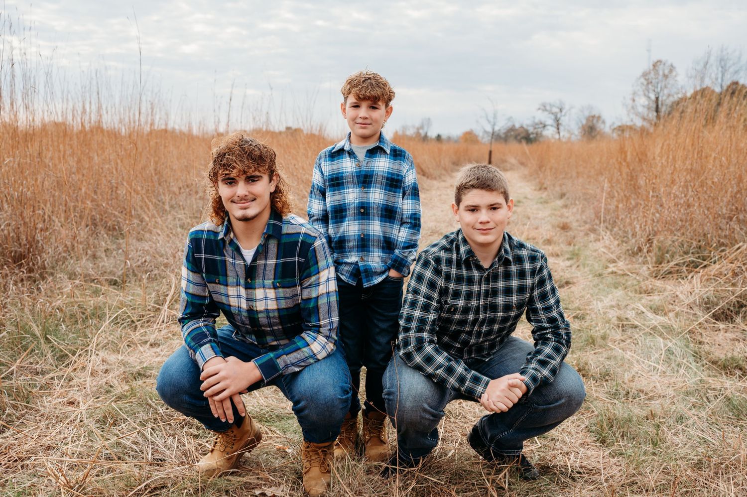 Three figures in plaid flannel shirts pose together in a field of tall brown grass on an autumn day.