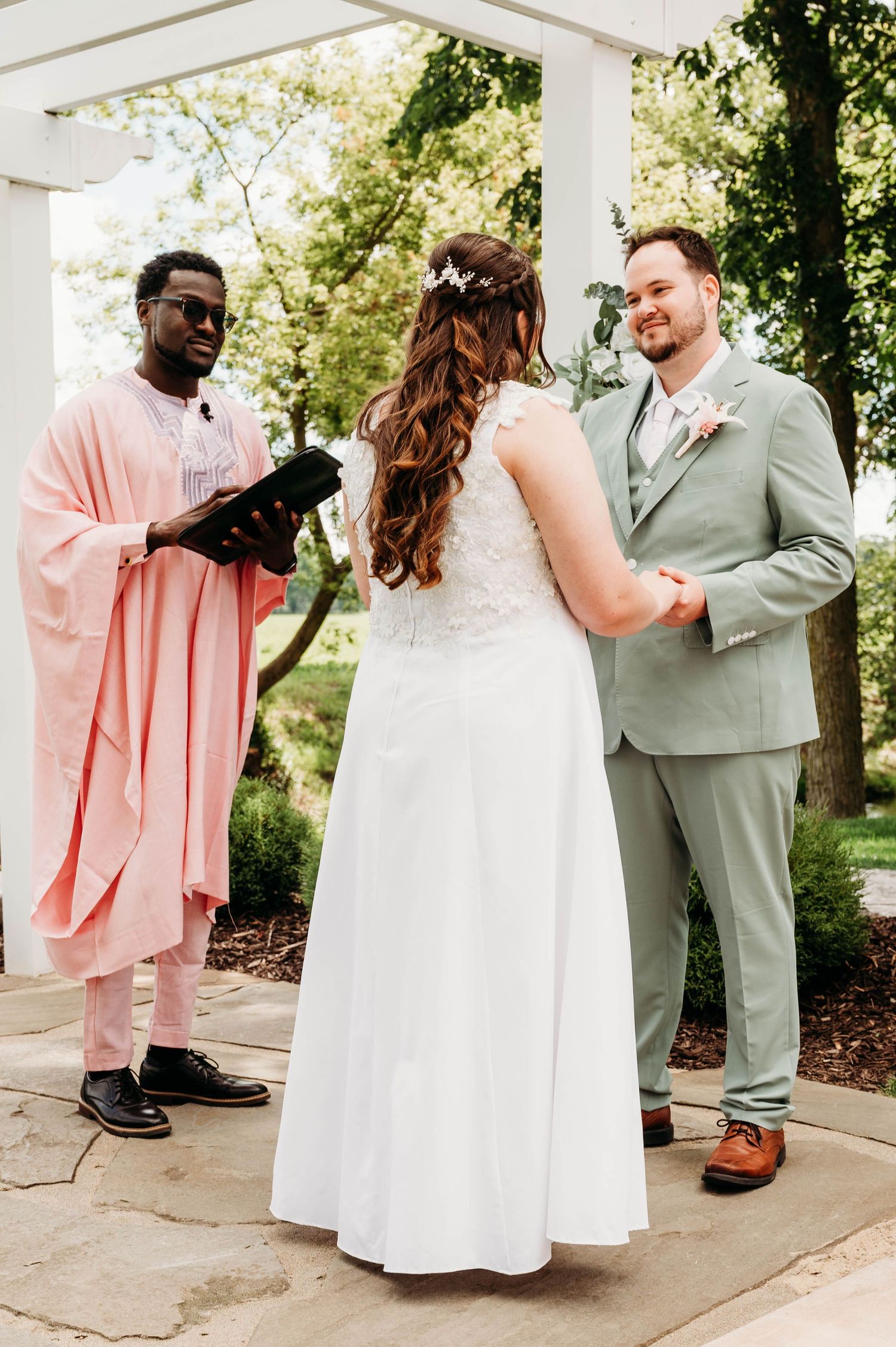 Bride and groom exchange vows outdoors under a white pergola, officiated by a man in a pink robe holding a book.